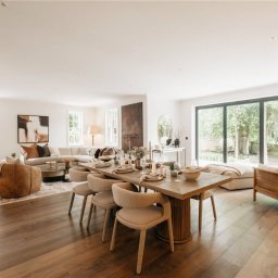 AW studio - Jasne, przestronne wnętrze domu z drewnianą podłogą, dużym oknem tarasowym, stołem z nakryciem i częścią wypoczynkową z beżową sofą narożną i fotelami.