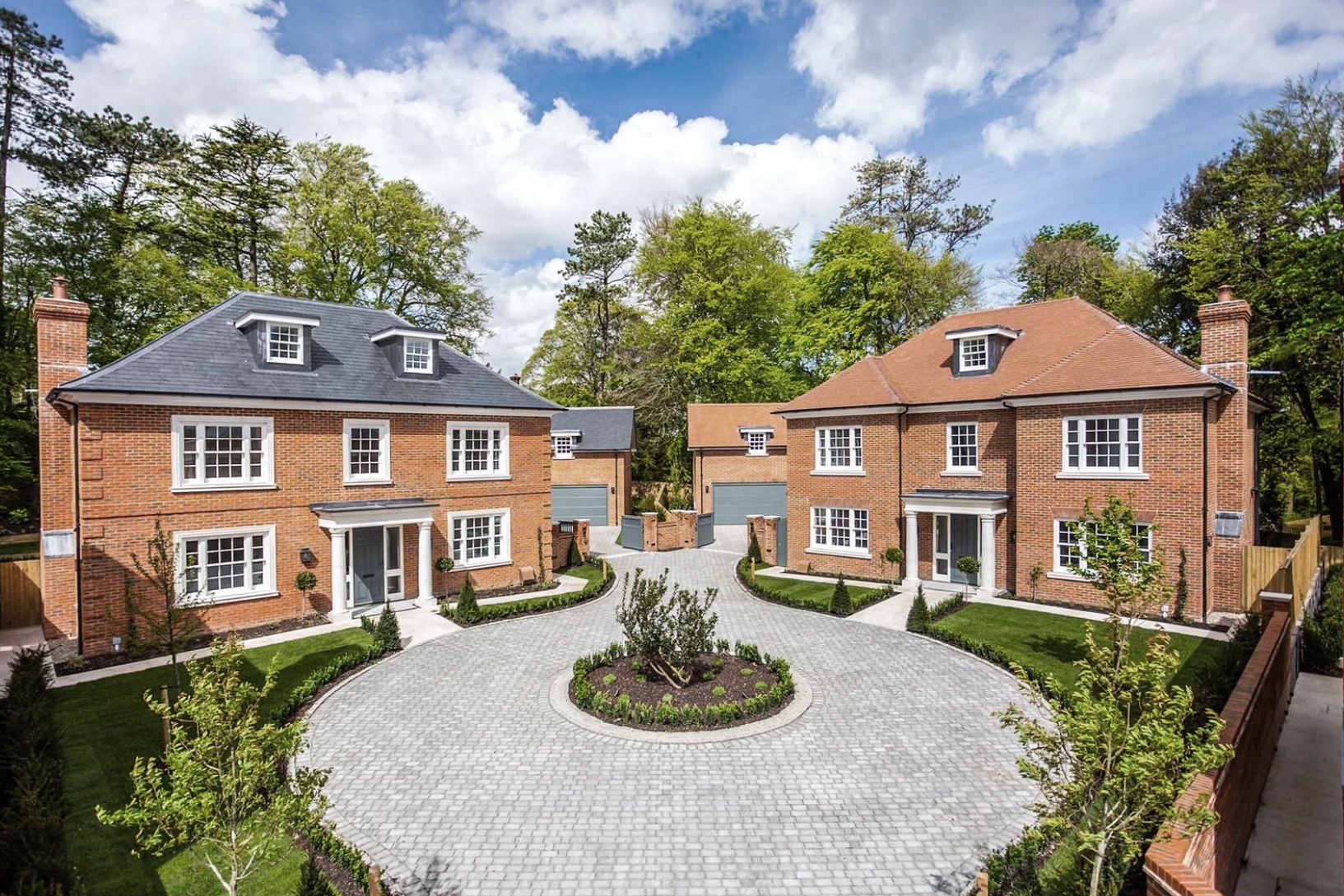 Dwa identyczne, dwupiętrowe domy z czerwonej cegły z szarymi dachami, symetrycznie usytuowane po obu stronach okrągłego podjazdu z kostki brukowej, otoczone zielenią i drzewami.