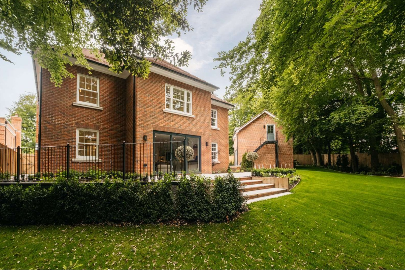 Dwupiętrowy dom z czerwonej cegły z dużymi oknami i przesuwnymi drzwiami tarasowymi, widok z zadbanego trawnika z drzewami i oddzielnym budynkiem gospodarczym z zewnętrznymi schodami.