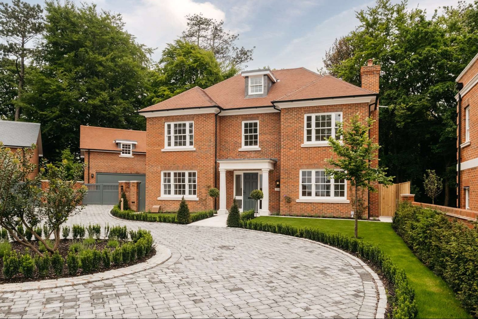 Dwukondygnacyjny dom z czerwonej cegły z kolumnowym wejściem i szarymi drzwiami, otoczony zadbanym trawnikiem i brukowanym podjazdem.
