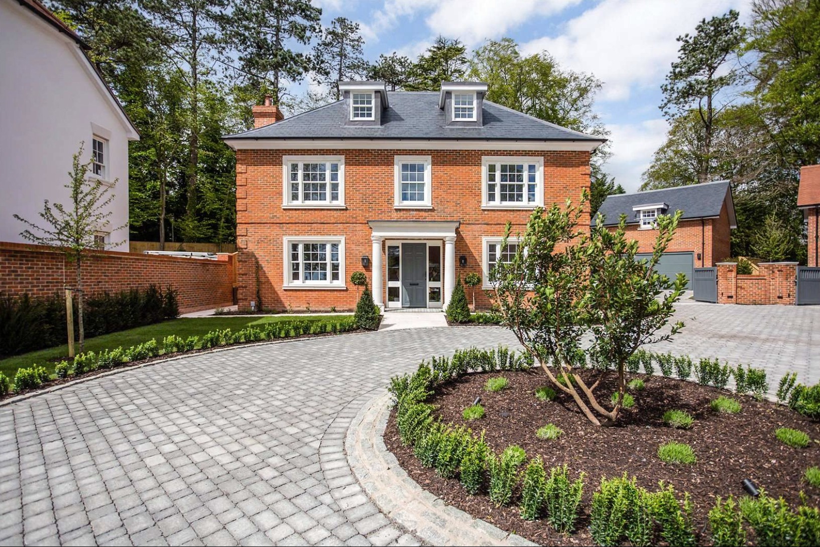 Dwukondygnacyjny dom z czerwonej cegły z szarymi oknami i drzwiami, kolumnami przy wejściu, szarym dachem z lukarnami, otoczony zielenią, z brukowanym podjazdem