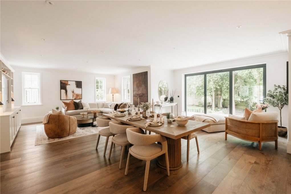 Jasne, przestronne wnętrze domu z drewnianą podłogą, dużym oknem tarasowym, stołem z nakryciem i częścią wypoczynkową z beżową sofą narożną i fotelami.