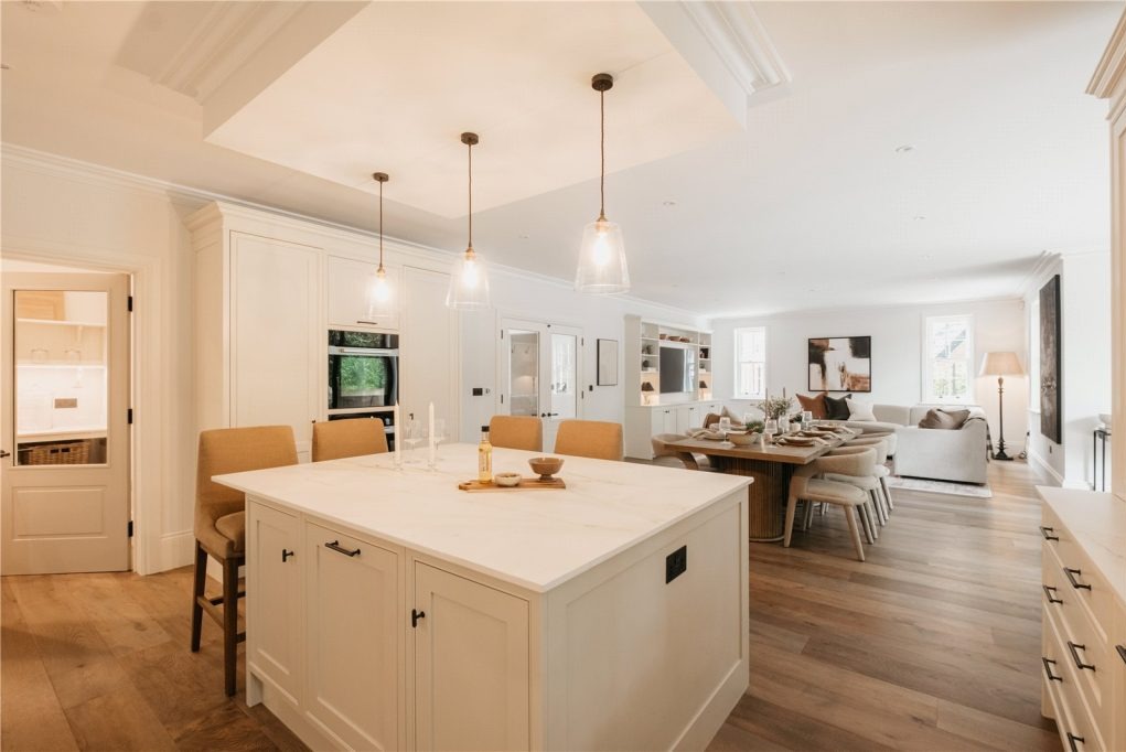 Jasna kuchnia otwarta na salon z wyspą kuchenną z blatem imitującym marmur, wiszące lampy ze szklanymi kloszami, stół jadalny z naturalnego drewna i wygodna sofa w tle.