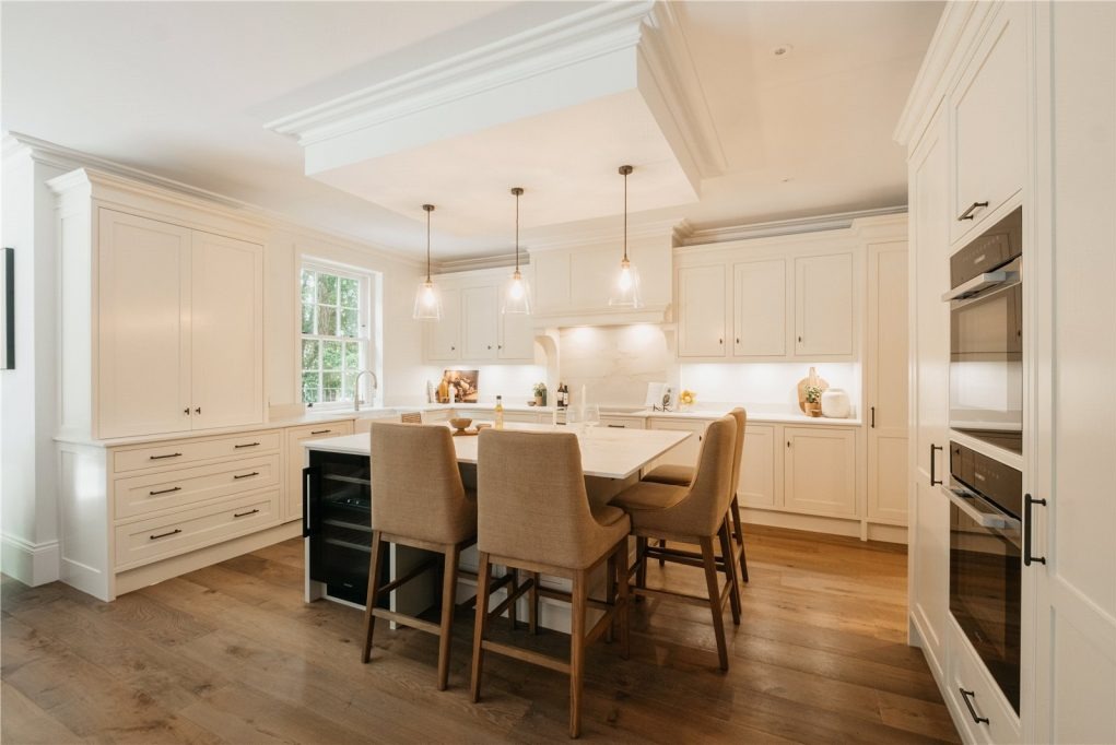 Jasna kuchnia z wyspą i czterema hokerami, drewniana podłoga, zabudowa kuchenna w stylu klasycznym, oświetlenie punktowe i wiszące lampy nad wyspą.