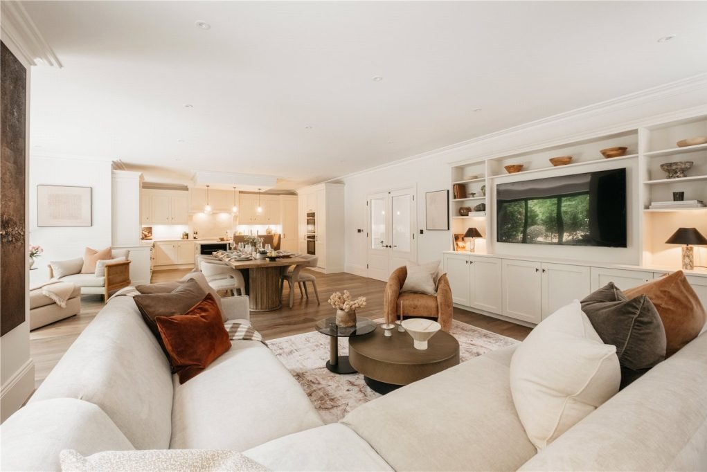 Przestronne wnętrze domu z otwartą kuchnią, jadalnią i salonem, utrzymane w jasnej kolorystyce z akcentami brązów i rudości, widoczne meble wypoczynkowe, stół z krzesłami oraz zabudowa telewizyjna.