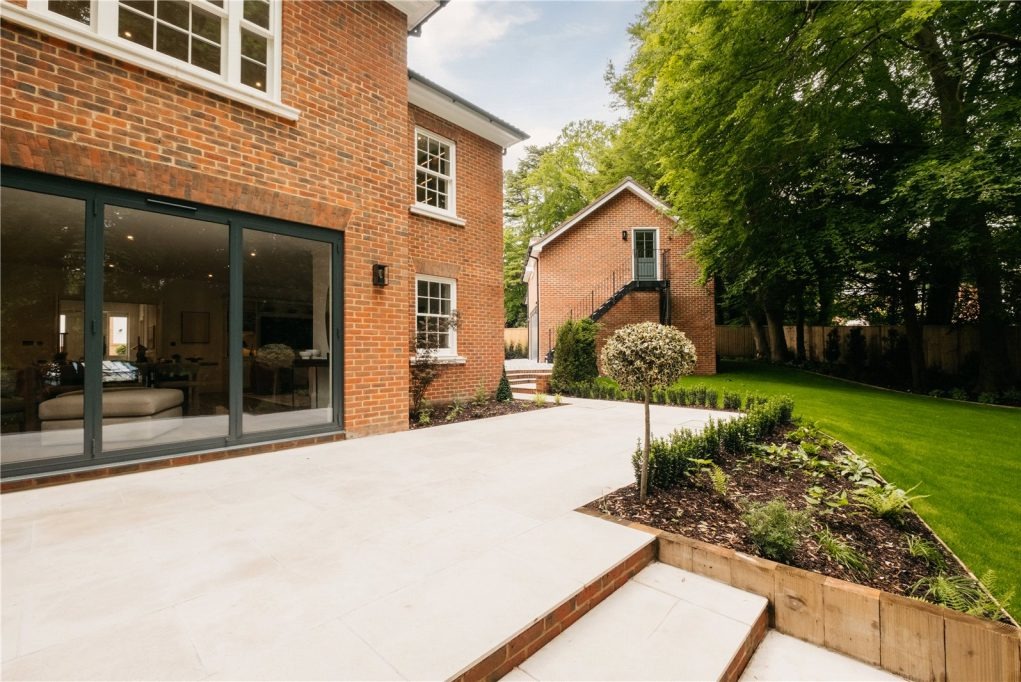 Widok na nowoczesny dom z cegły z rozległym tarasem z jasnych płyt, przesuwnymi szklanymi drzwiami i zadbanym ogrodem z równo przystrzyżonym trawnikiem i dekoracyjnymi krzewami; w tle widoczny...