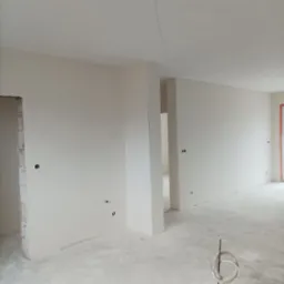 Wnętrze w stanie deweloperskim po wykonaniu prac wykończeniowych ścian i sufitu, widoczne gładkie powierzchnie przygotowane do malowania, wystające przewody elektryczne i rury instalacyjne.