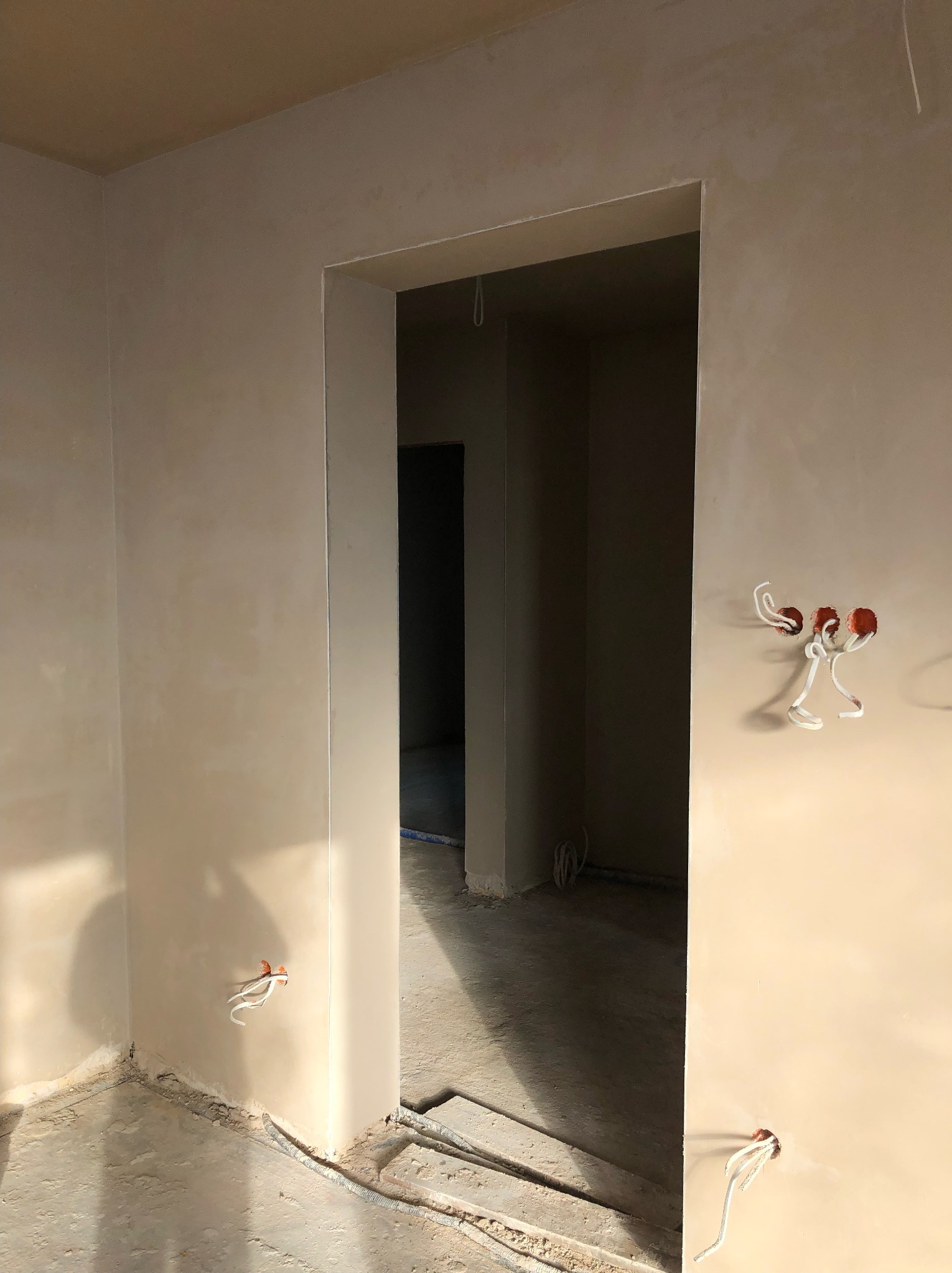 Wnętrze w trakcie prac wykończeniowych: ściany pokryte świeżo nałożoną, jasną warstwą wygładzającą, widoczne wystające przewody elektryczne, surowy beton na podłodze i fragment przejścia...