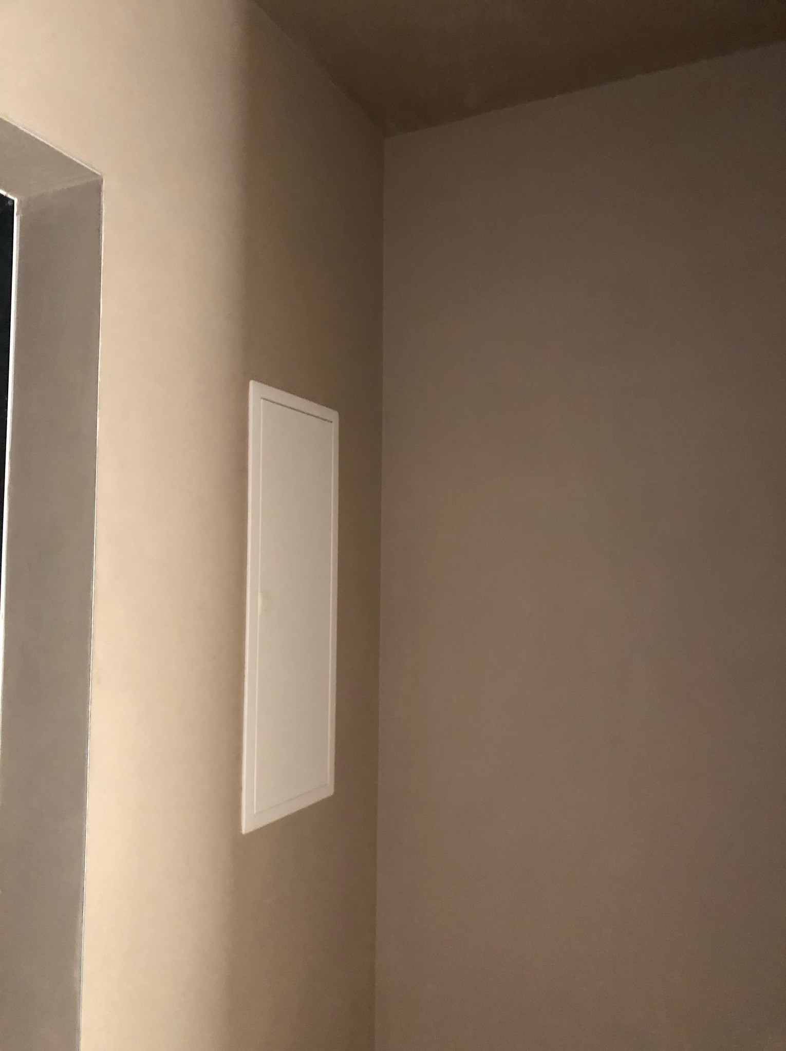 Fragment wykończonego wnętrza: gładka, beżowa ściana z zamontowaną białą, prostokątną klapą rewizyjną. Widoczne łączenie ściany z sufitem oraz fragment ościeżnicy po lewej stronie.