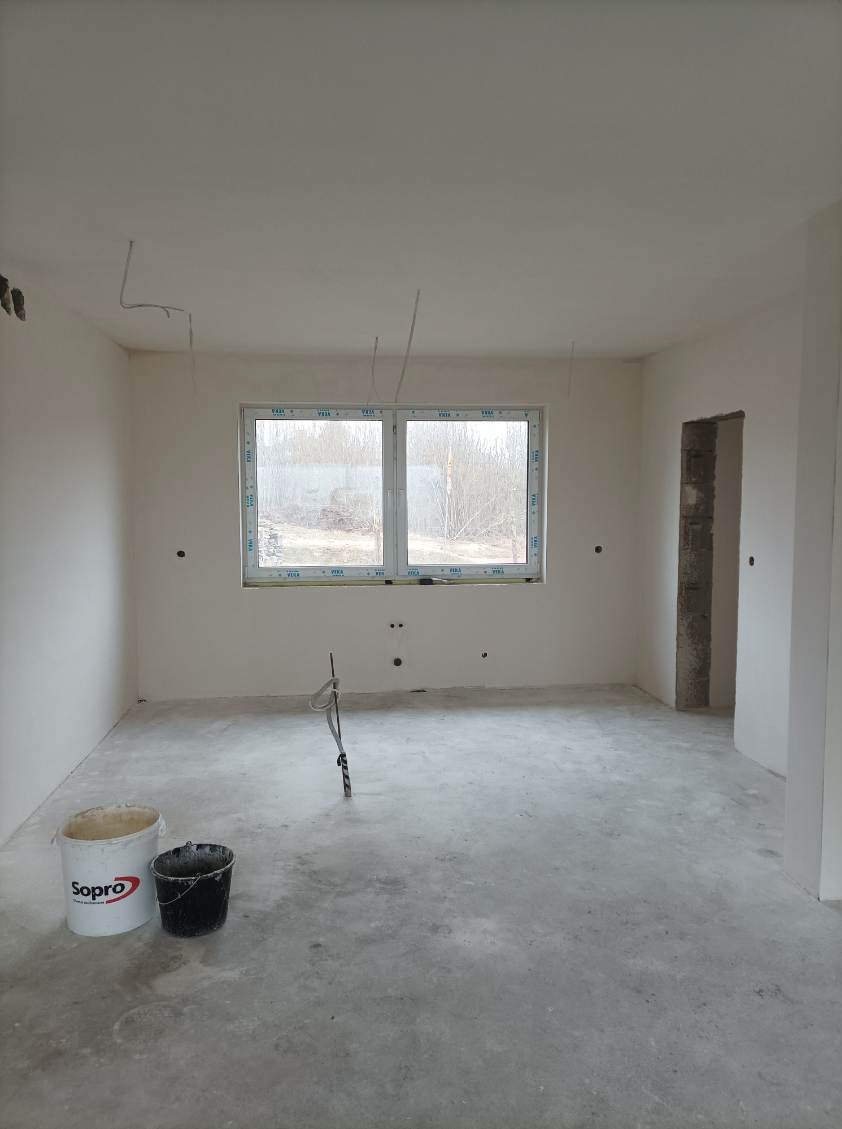 Surowe wnętrze pokoju po wstępnych pracach wykończeniowych, z widocznymi przewodami elektrycznymi, oknem na zewnątrz, wiadrami z resztkami materiałów budowlanych i świeżo położoną, szarą wylewką...