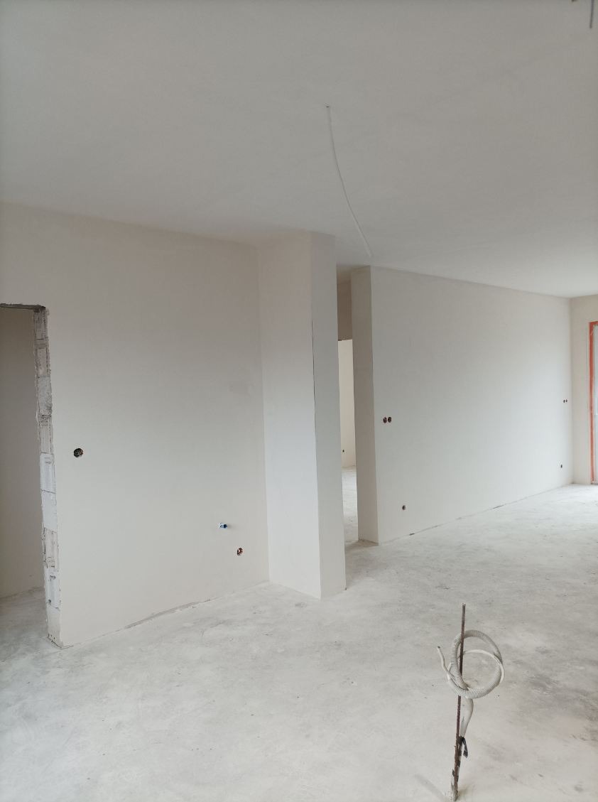 Wnętrze w stanie deweloperskim po wykonaniu prac wykończeniowych ścian i sufitu, widoczne gładkie powierzchnie przygotowane do malowania, wystające przewody elektryczne i rury instalacyjne.