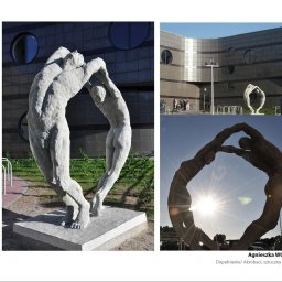 ARCHITEKTURA I RZEŹBA AGNIESZKA WOLSKA-CIELIŃSKA - Kompozycja z dwóch rzeźb z jasnego kamienia, przedstawiających postacie w dynamicznej pozie, na tle nowoczesnego budynku z okrągłymi oknami i fragmentu parkingu w słoneczny dzień.
