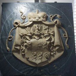 ARCHITEKTURA I RZEŹBA AGNIESZKA WOLSKA-CIELIŃSKA - Gipsowy herb z koroną i wężami, umieszczony na ciemnym tle, w pracowni artystycznej z widocznymi narzędziami i elementami wyposażenia.