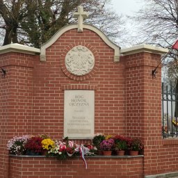 ARCHITEKTURA I RZEŹBA AGNIESZKA WOLSKA-CIELIŃSKA - Murowane ogrodzenie cmentarza z cegły z polskim godłem i napisem patriotycznym, udekorowane kwiatami. Flagi Polski po bokach.