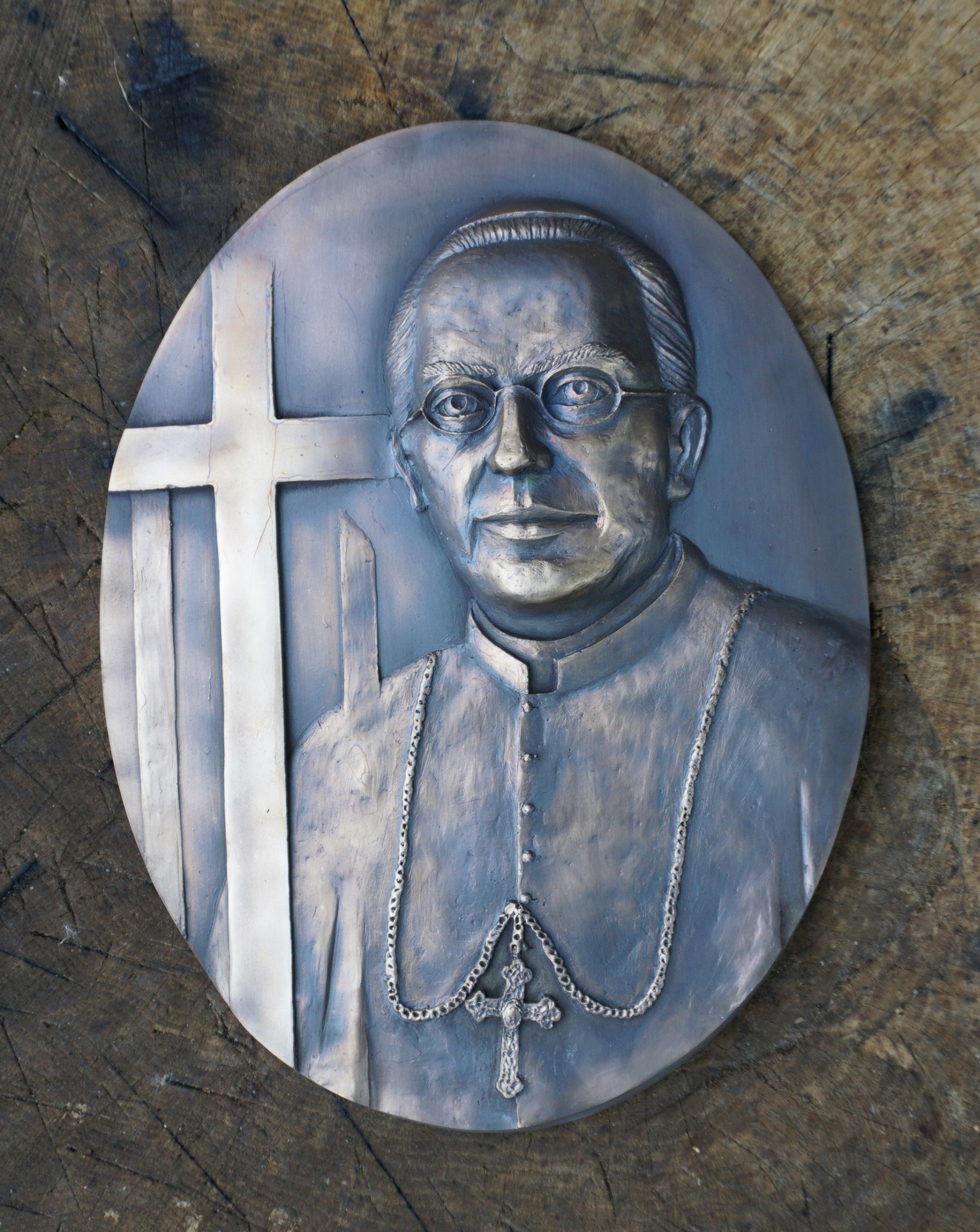 Metalowa płaskorzeźba przedstawiająca portret biskupa w okularach, z krzyżem w tle, umieszczona na drewnianym pniu.
