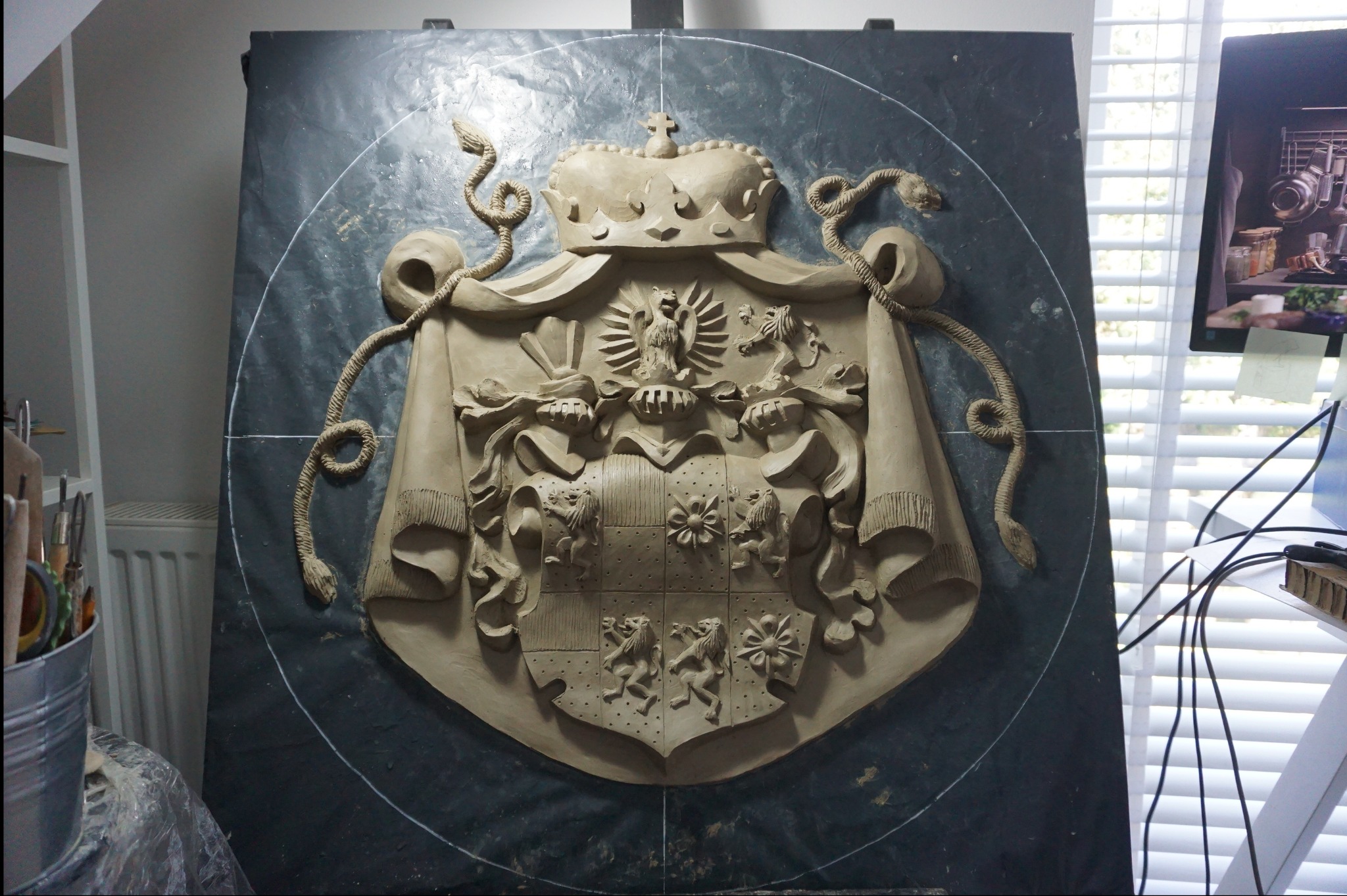 Gipsowy herb z koroną i wężami, umieszczony na ciemnym tle, w pracowni artystycznej z widocznymi narzędziami i elementami wyposażenia.