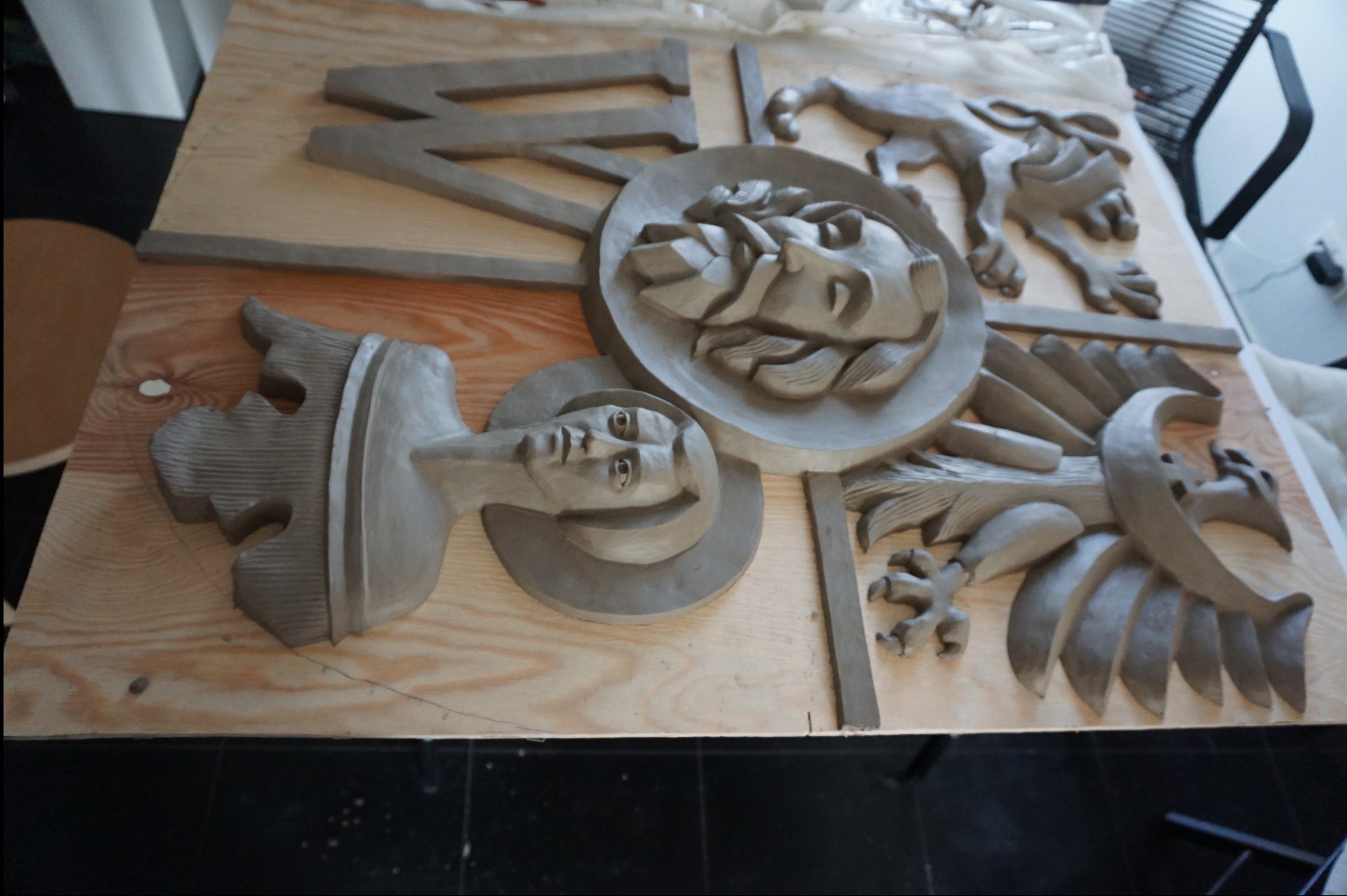 Gliniana płaskorzeźba z motywami religijnymi na drewnianej płycie: portrety i symboliczne zwierzęta. Praca w toku, detale w trakcie modelowania, surowy materiał.