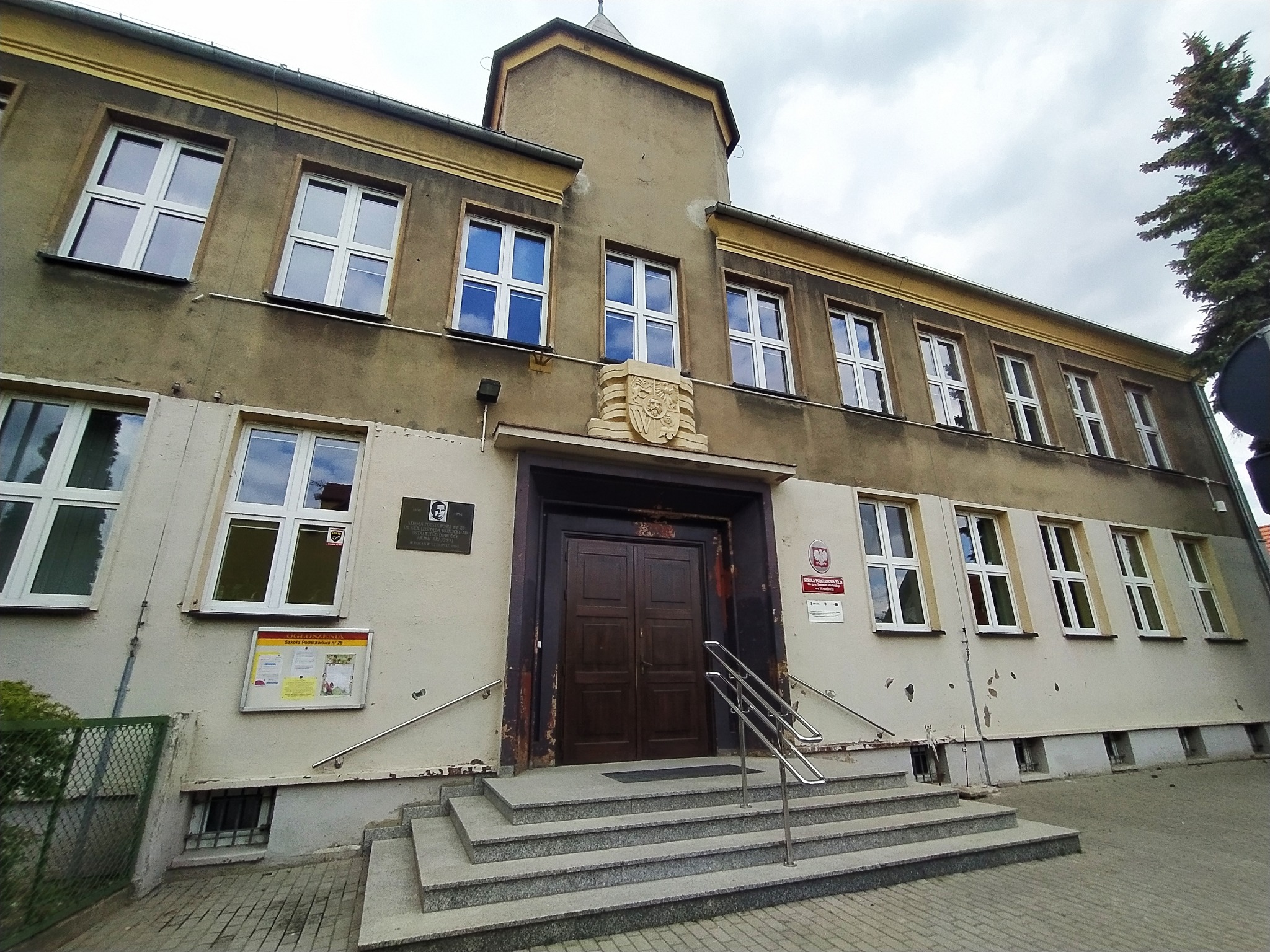 Fasada dwukondygnacyjnego budynku szkolnego z brązowymi drzwiami wejściowymi i schodami. Na elewacji tablice informacyjne i godło. Widok z dołu, pochmurne niebo.