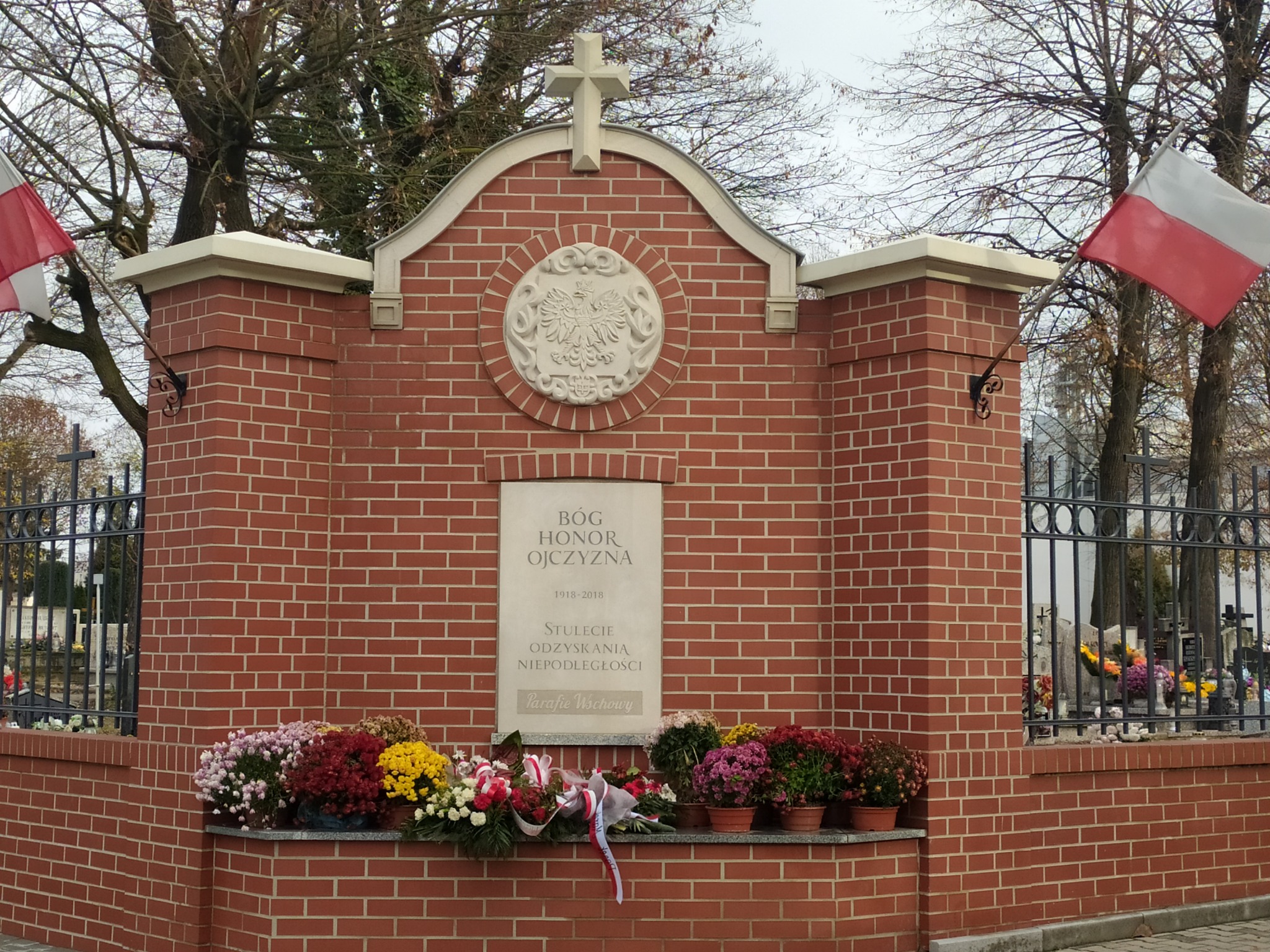 Murowane ogrodzenie cmentarza z cegły z polskim godłem i napisem patriotycznym, udekorowane kwiatami. Flagi Polski po bokach.