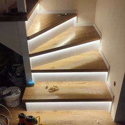 Mikalai Voitau - Drewniane schody z subtelnym oświetleniem LED pod stopniami, narzędzia pracy na podłodze. Ciepłe światło podkreśla naturalną teksturę drewna.