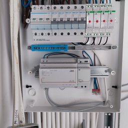 Mikalai Voitau - Rozdzielnica elektryczna z widocznymi zabezpieczeniami, przewodami i elementami sterującymi, zamontowana na białej ścianie. Widoczne kable i skrzynka przyłączeniowa.