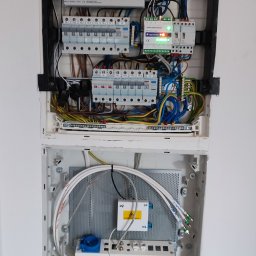 Mikalai Voitau - Otwarta skrzynka elektryczna z bezpiecznikami, przewodami i modułem światłowodowym. Widoczne kable w różnych kolorach i elementy instalacji.