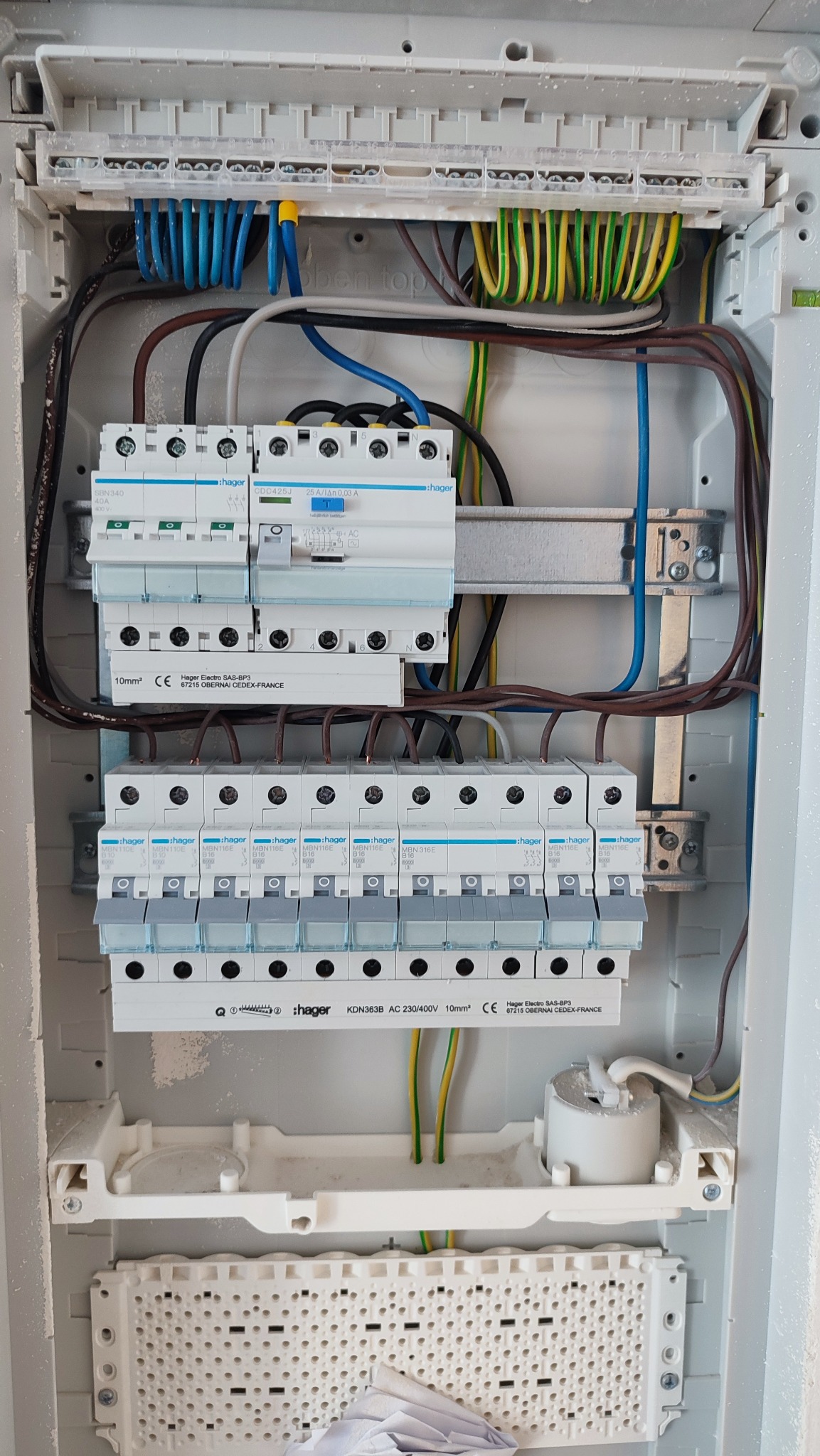 Rozdzielnica elektryczna Hager z zainstalowanymi bezpiecznikami i okablowaniem. Widoczne przewody w różnych kolorach i elementy montażowe. Biała obudowa rozdzielnicy.