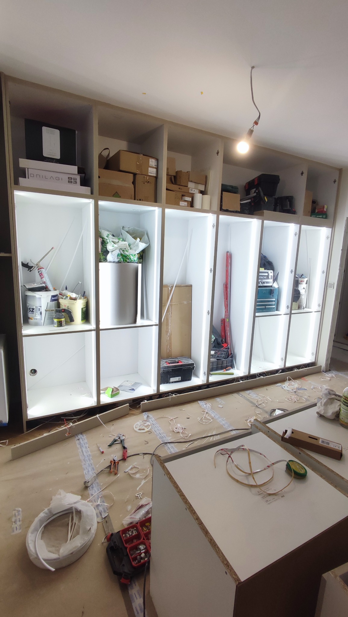 Montaż szafy wnękowej z oświetleniem LED. Widoczne narzędzia i akcesoria montażowe. Szafa w trakcie instalacji, z otwartymi półkami i elementami wyposażenia.