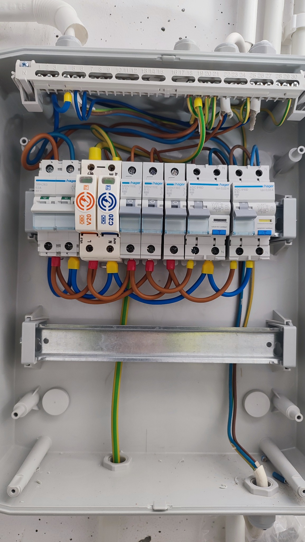 Rozdzielnica elektryczna Hager z zainstalowanymi bezpiecznikami i przewodami o różnych kolorach, zamontowana w szarej obudowie. Widoczne elementy instalacji elektrycznej.