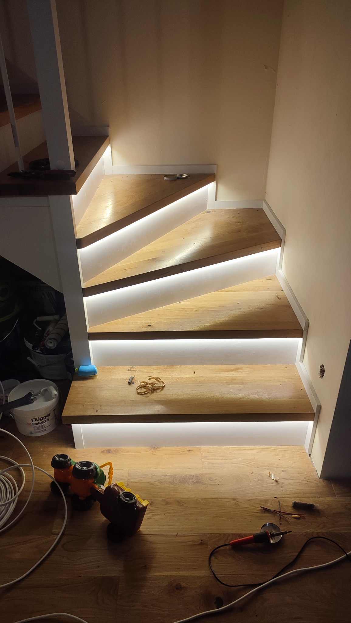 Drewniane schody z subtelnym oświetleniem LED pod stopniami, narzędzia pracy na podłodze. Ciepłe światło podkreśla naturalną teksturę drewna.