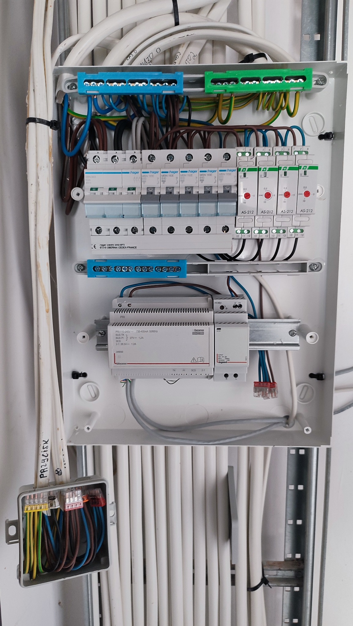 Rozdzielnica elektryczna z widocznymi zabezpieczeniami, przewodami i elementami sterującymi, zamontowana na białej ścianie. Widoczne kable i skrzynka przyłączeniowa.