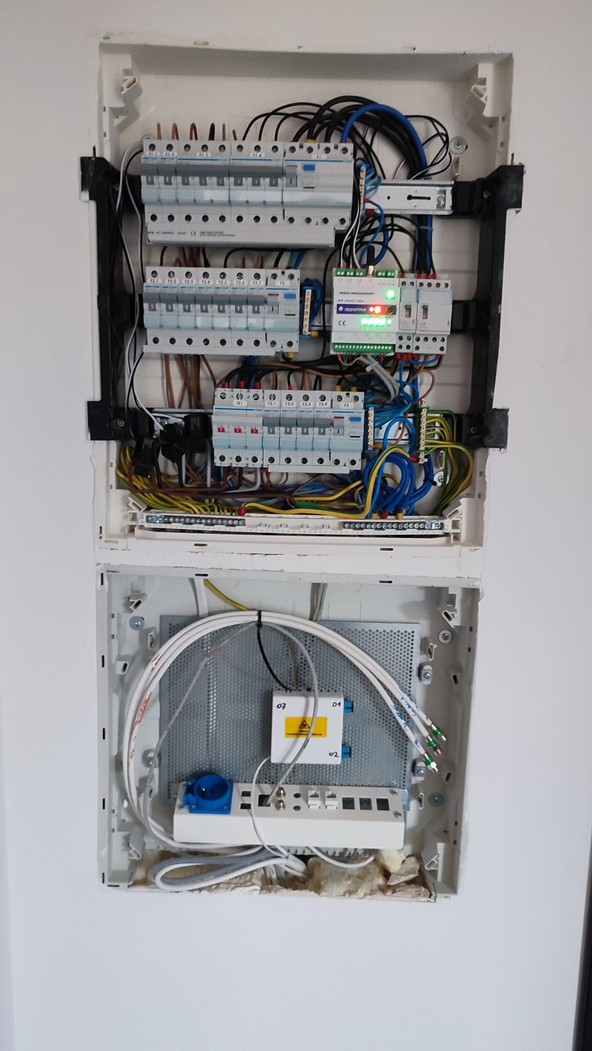 Otwarta skrzynka elektryczna z bezpiecznikami, przewodami i modułem światłowodowym. Widoczne kable w różnych kolorach i elementy instalacji.