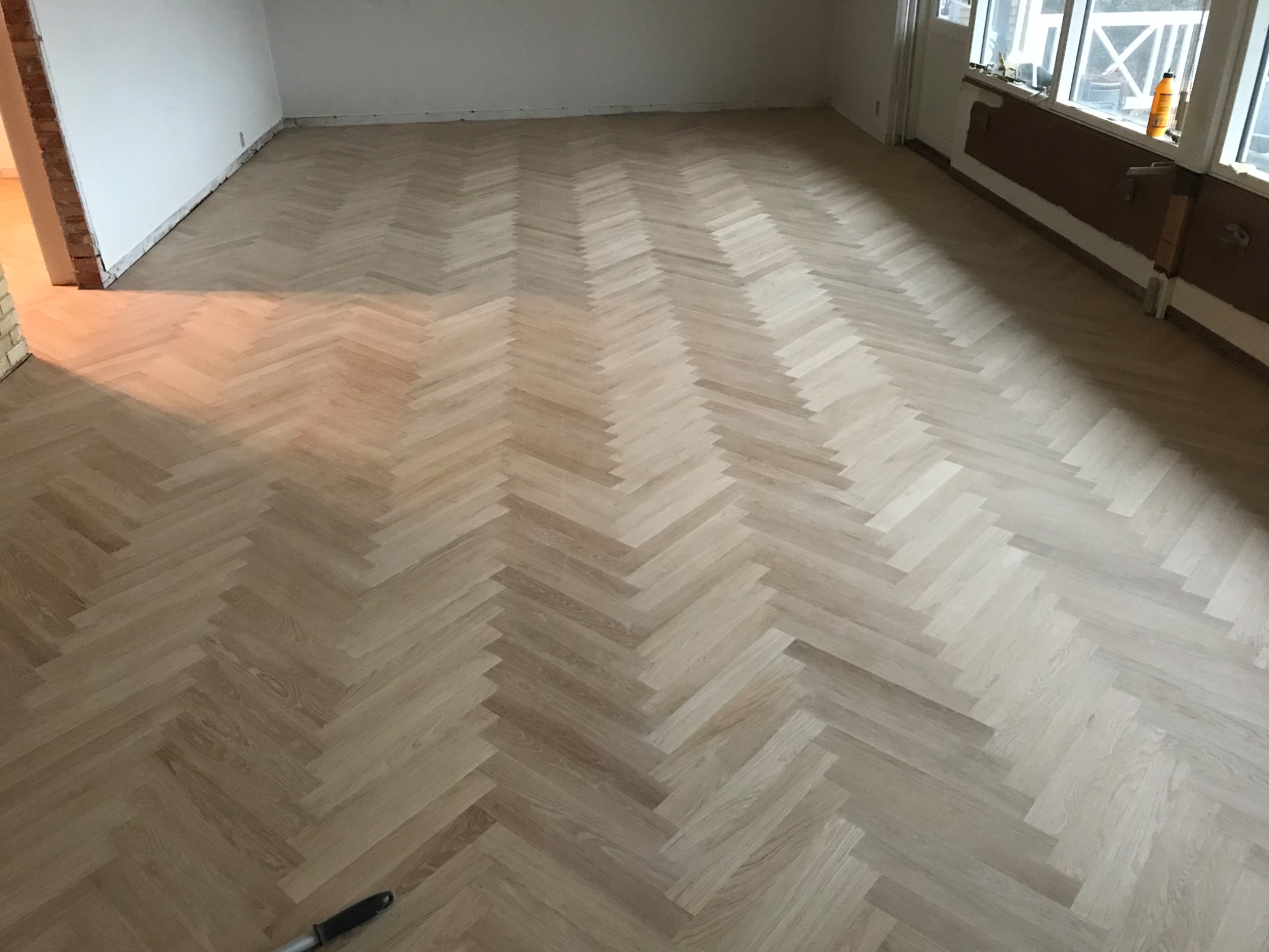 Jasny parkiet ułożony we wzór jodełki w przestronnym pokoju, widoczne niedokończone wykończenia ścian i listew przypodłogowych.