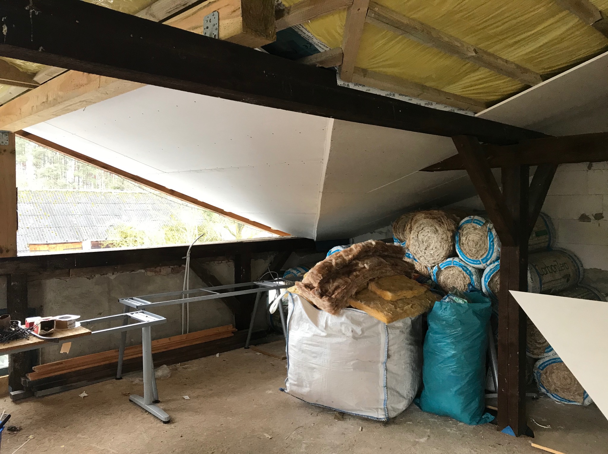 Poddasze w trakcie adaptacji: częściowo zamontowana płyta karton-gips na skosach, widoczne belki konstrukcyjne, izolacja termiczna w rolkach i workach, metalowe nogi stołu, okno dachowe z widokiem...