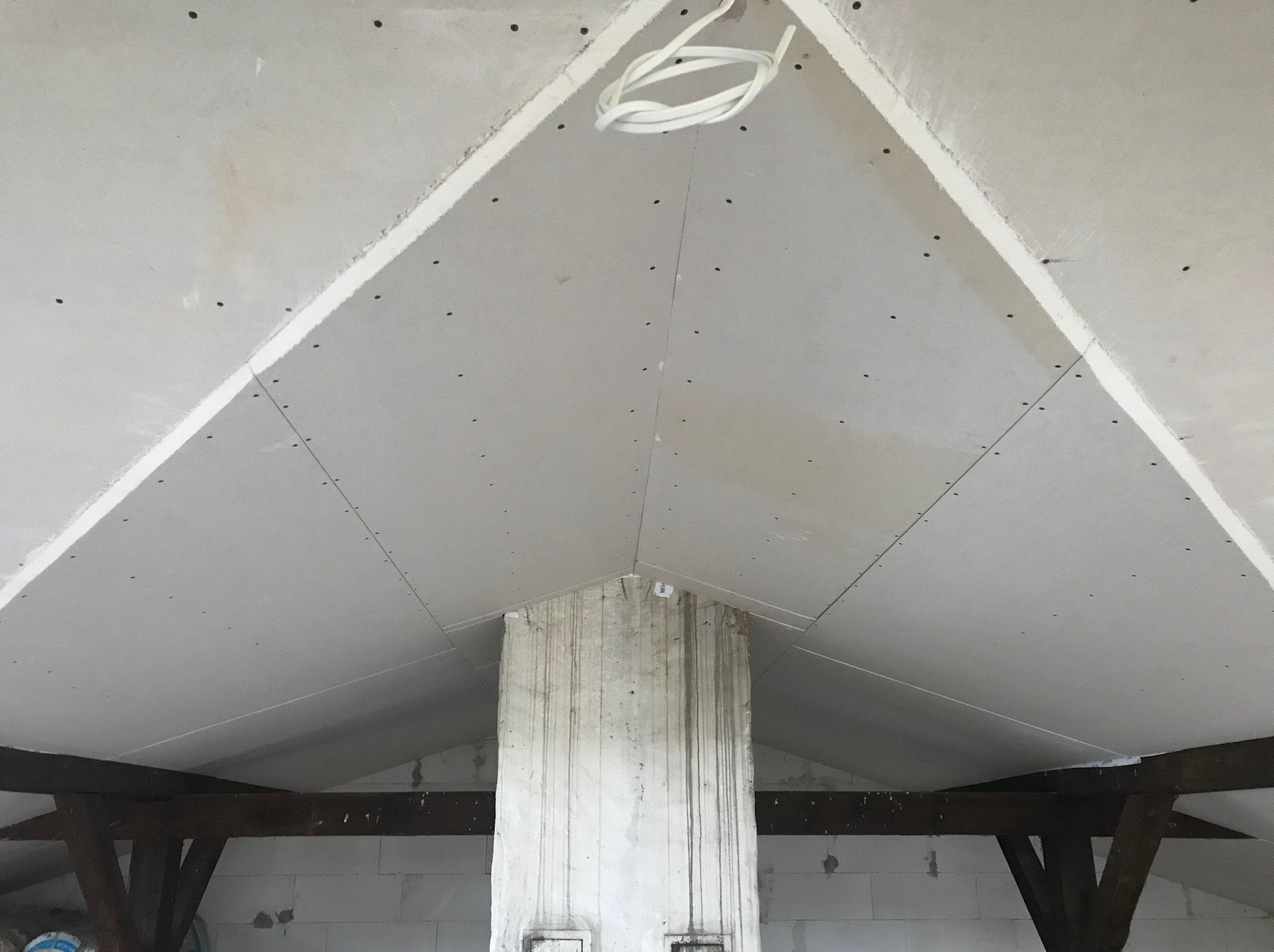 Skośny sufit poddasza z płyt kartonowo-gipsowych, widoczne łączenia płyt, przewody elektryczne wystające z sufitu, drewniana konstrukcja nośna i fragment ściany z bloczków.