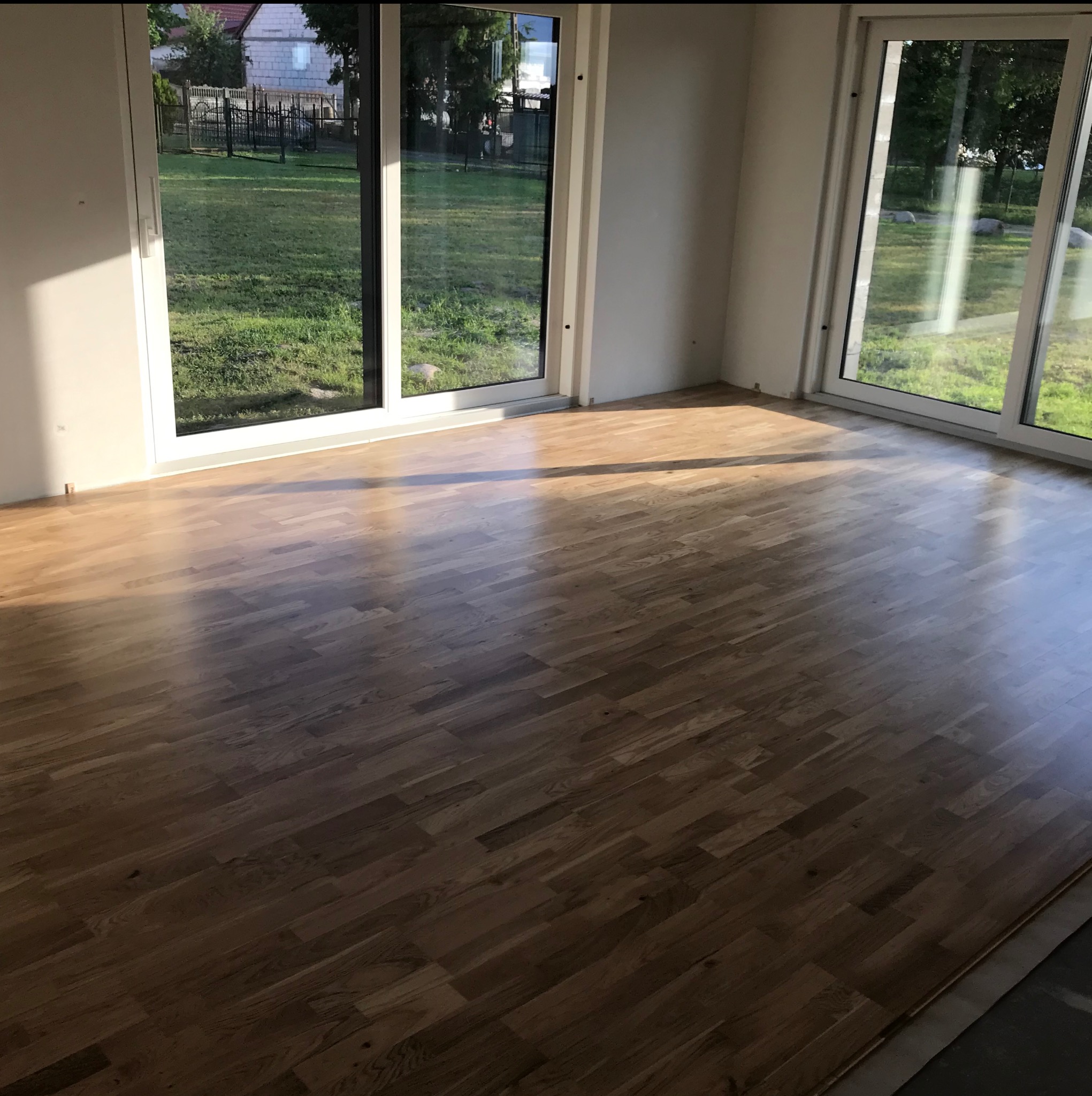 Ułożony parkiet w jasnym pokoju z dużymi oknami wychodzącymi na ogród, widoczne niedociągnięcia przy ścianie.