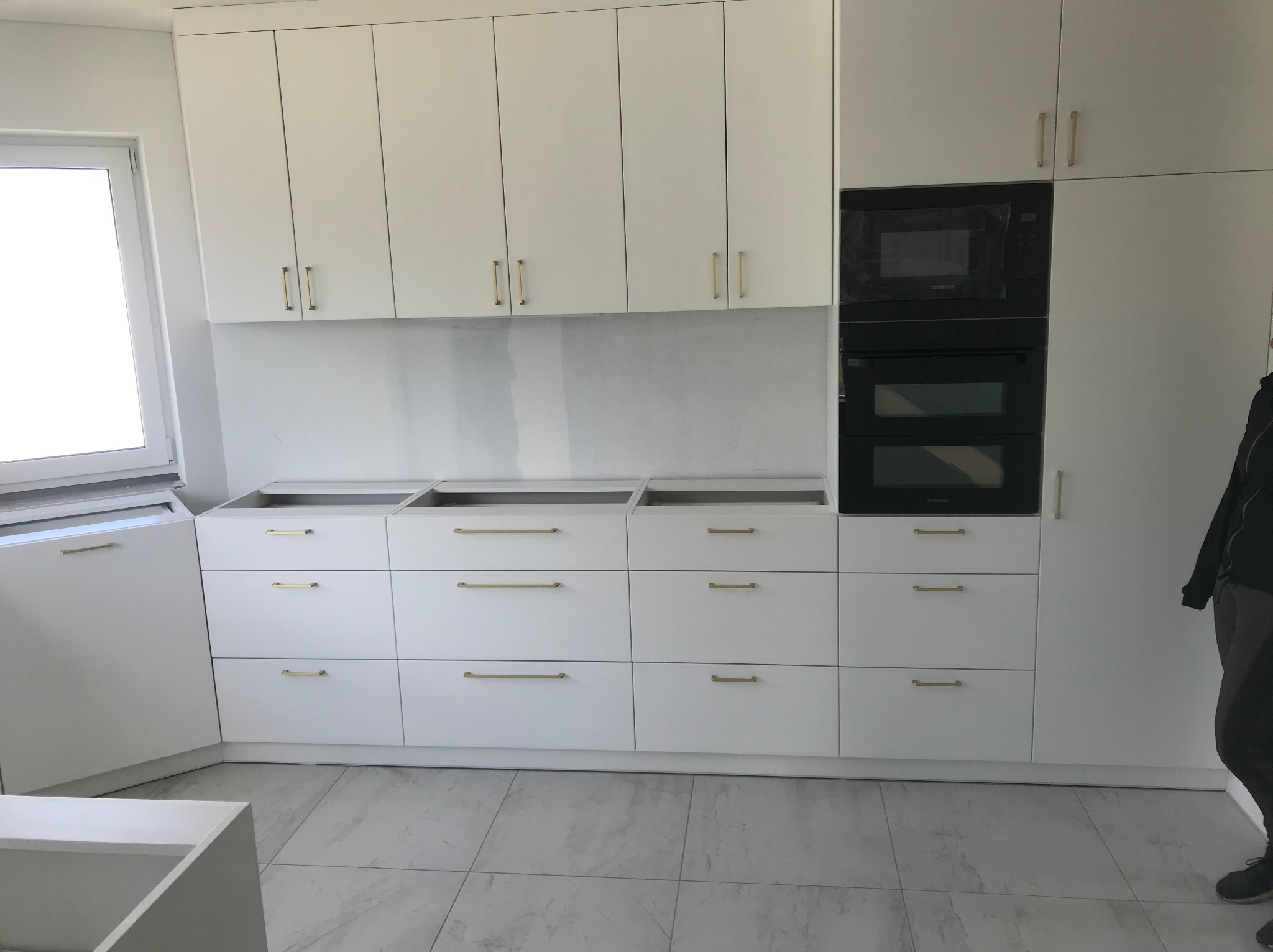 Biała zabudowa kuchenna z czarnymi piekarnikami w trakcie montażu, widoczne szuflady bez blatów i osoba w tle.