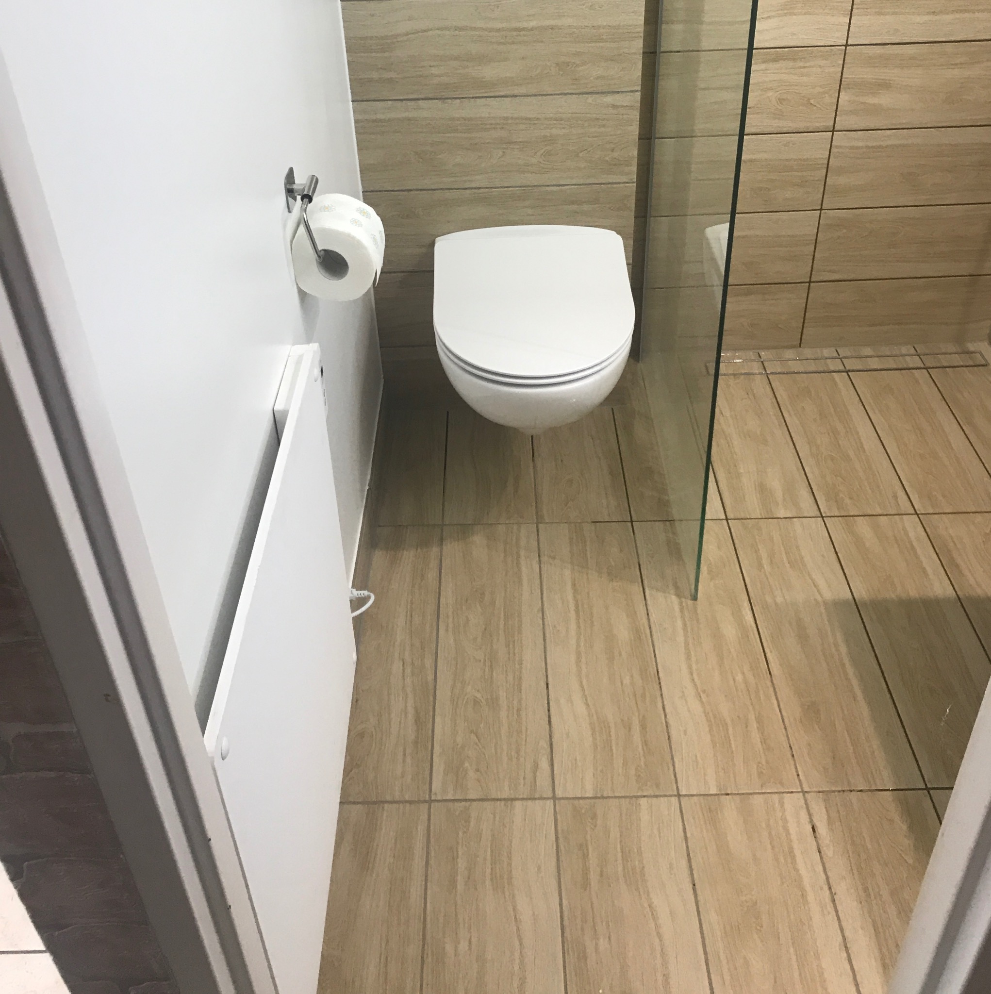 Wykończona łazienka z toaletą wiszącą, płytkami imitującymi drewno na ścianach i podłodze, kabiną prysznicową ze szklaną ścianką oraz grzejnikiem elektrycznym zamontowanym na ścianie obok rolki...