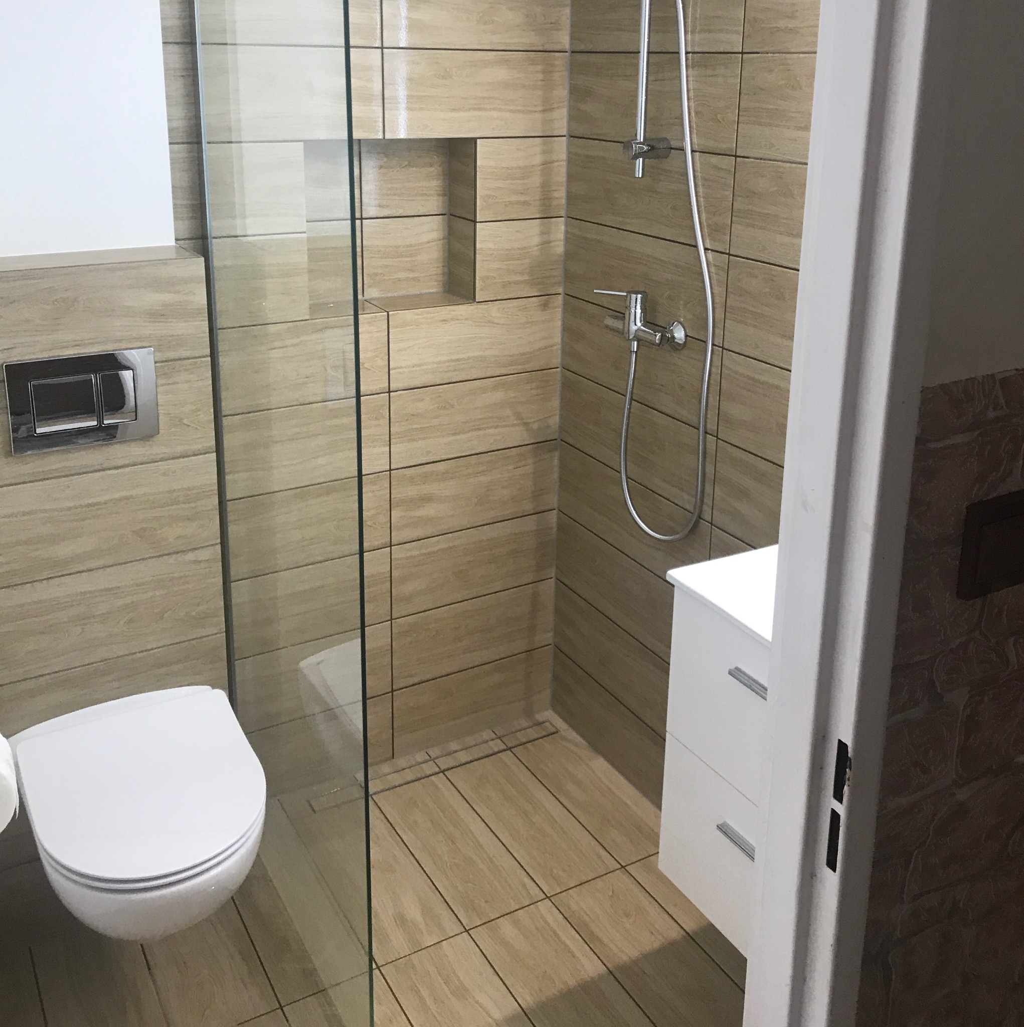 Nowoczesna łazienka z kabiną prysznicową typu walk-in, wykończona płytkami imitującymi drewno, z podtynkową spłuczką WC i białą szafką pod umywalką z dwoma szufladami.