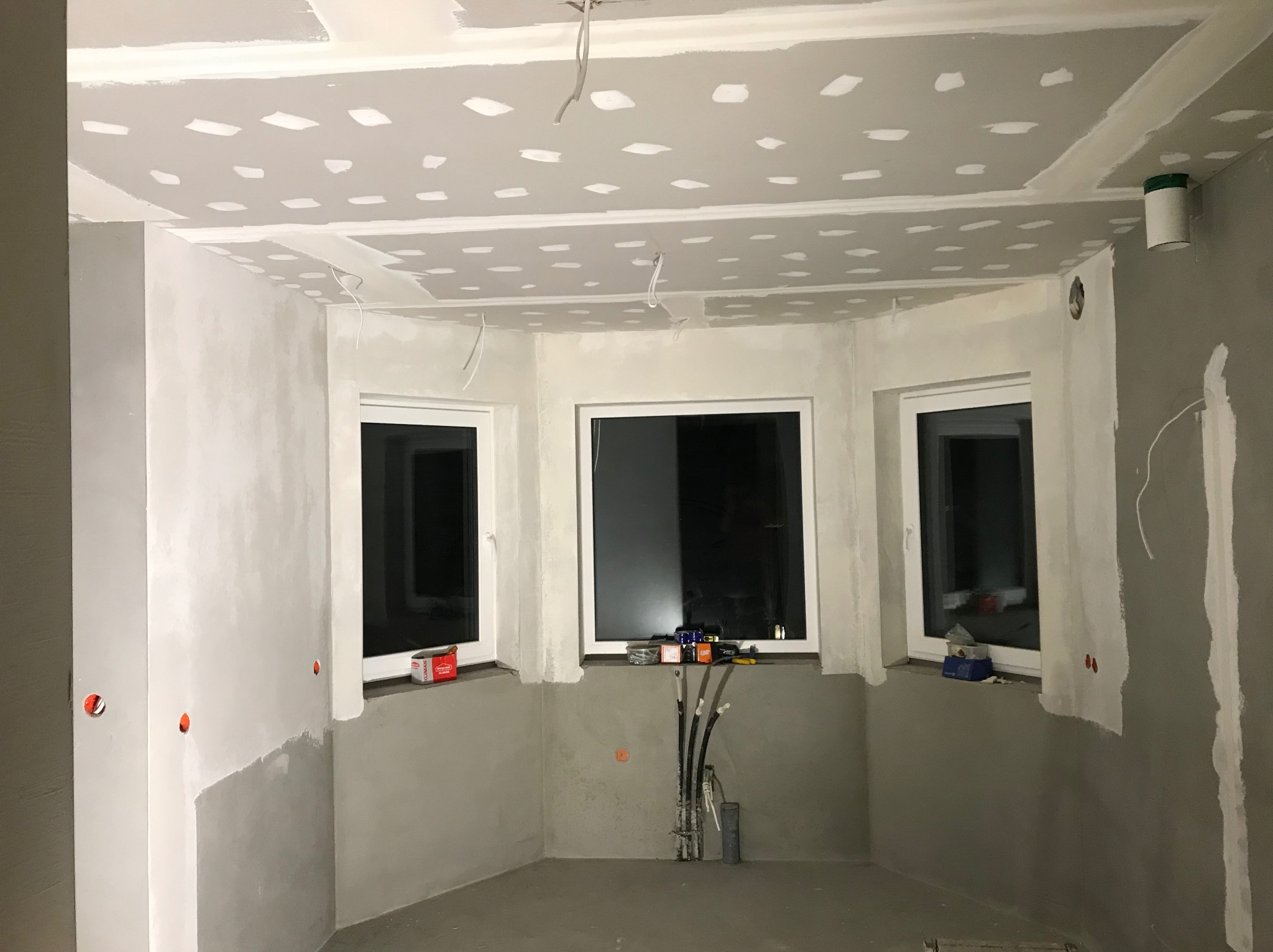 Wnętrze pokoju z trzema oknami w trakcie montażu płyt karton gips na suficie i ścianach, widoczne szpachlowane łączenia płyt, wystające przewody elektryczne, narzędzia na parapetach.