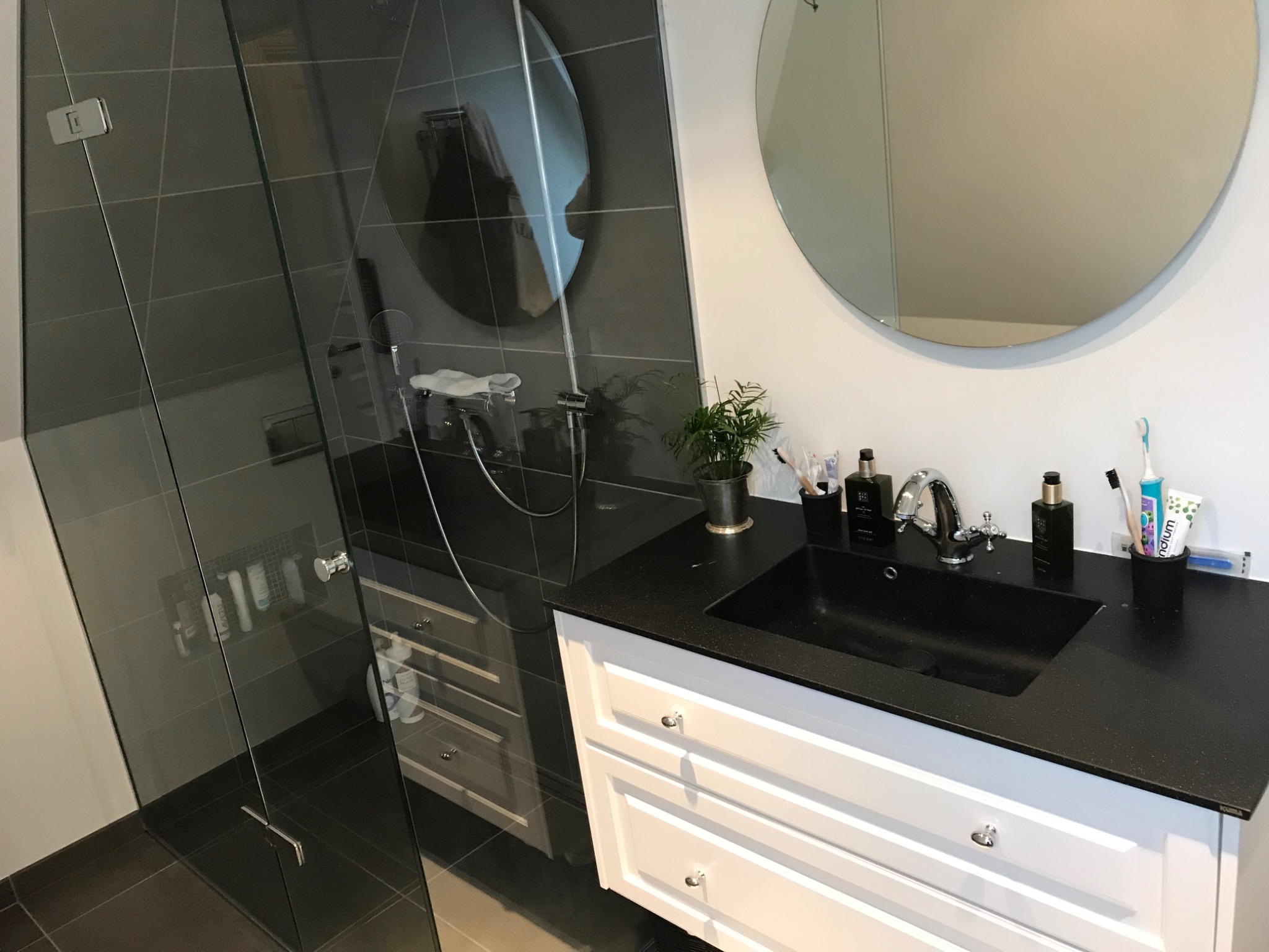 Nowoczesna łazienka z czarną umywalką nablatową, białą szafką z frezowanymi frontami, okrągłym lustrem i kabiną prysznicową z szarymi płytkami.