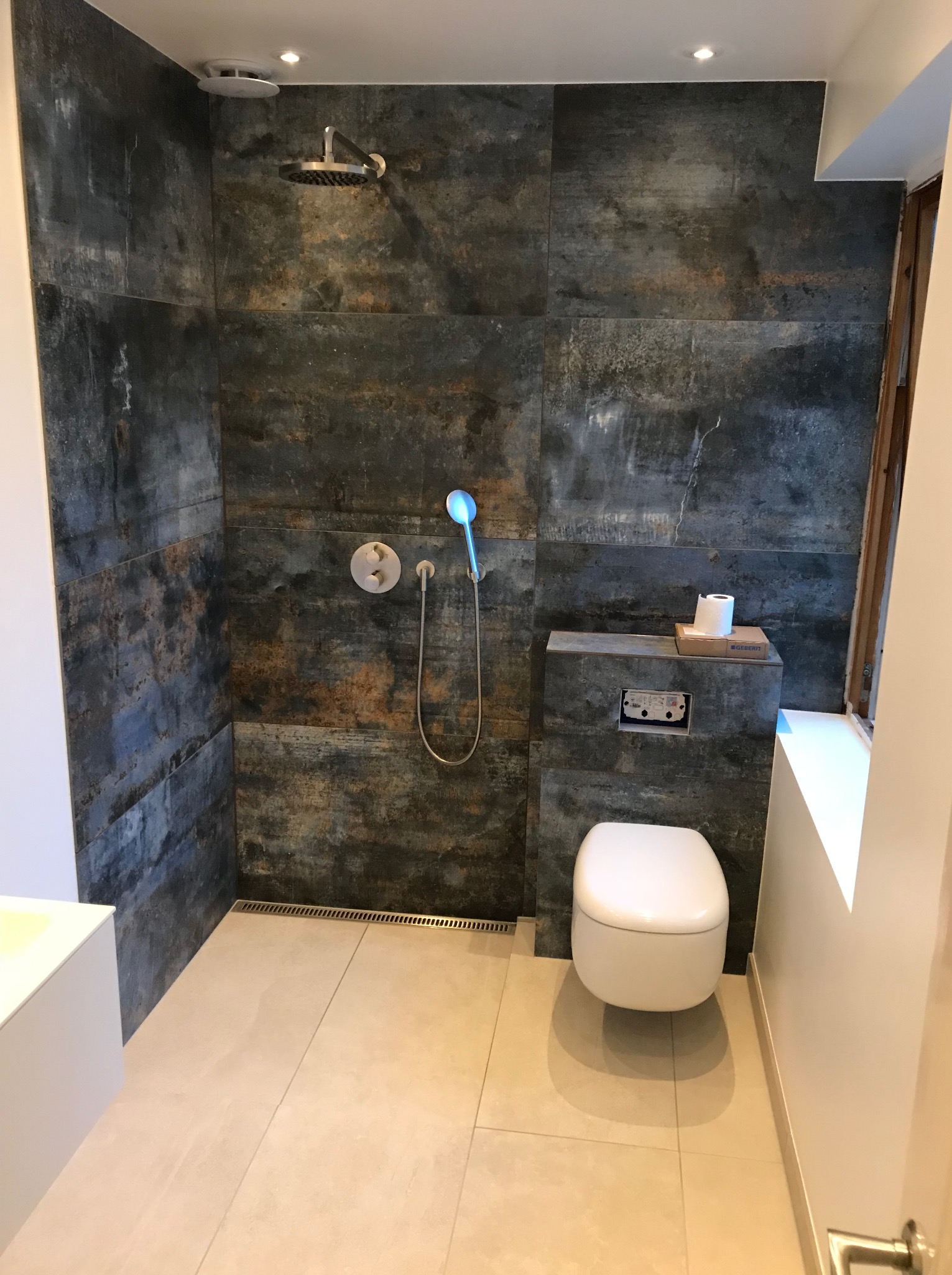 Nowoczesna łazienka z kabiną prysznicową wyłożoną płytkami imitującymi rdzę, wiszącą toaletą i odpływem liniowym w podłodze.