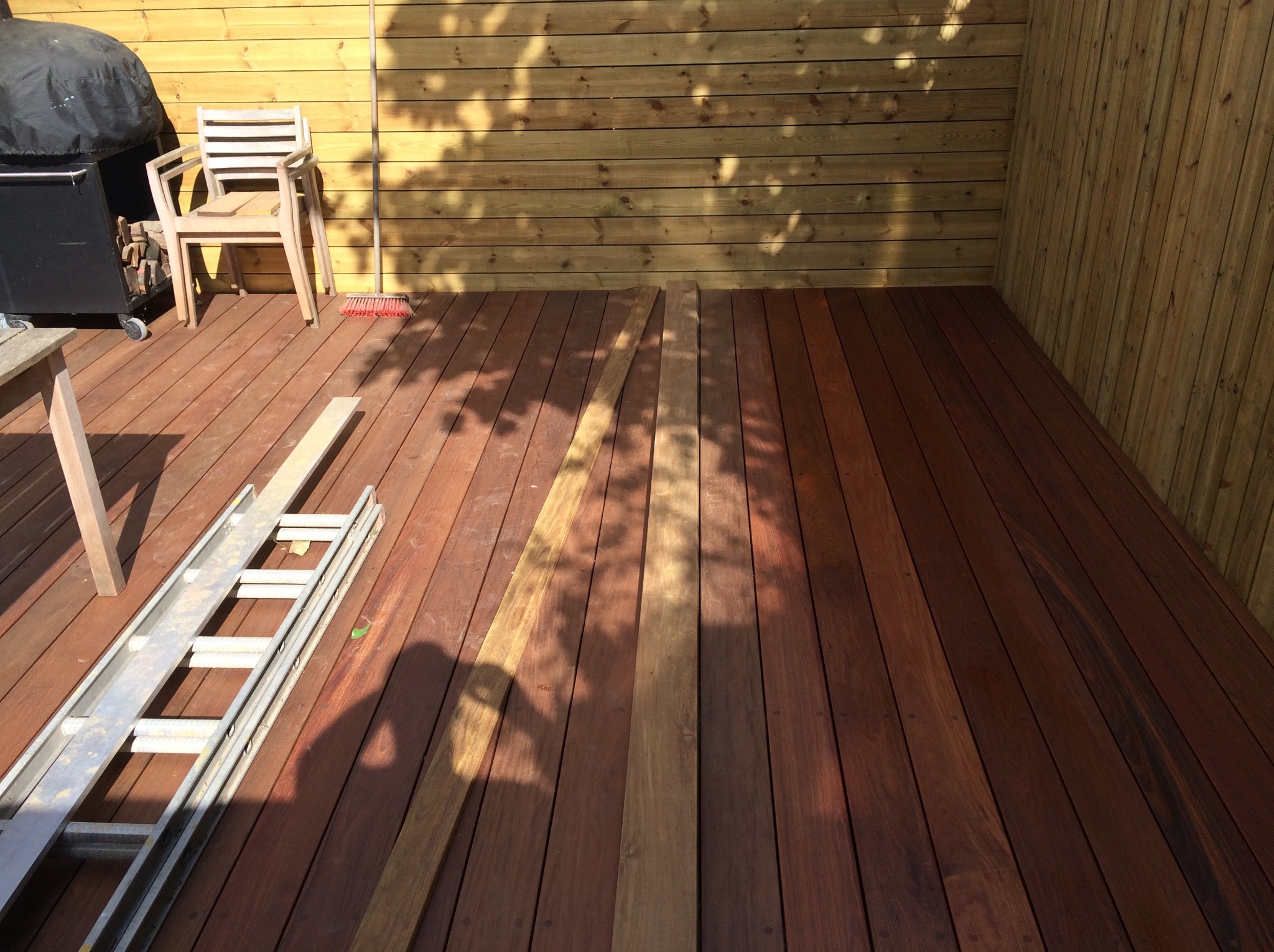 Nowa drewniana podłoga tarasu w trakcie montażu, deski w różnych odcieniach brązu, drabina aluminiowa leży na podłodze, drewniana ściana w tle, cień drzew na ścianie.