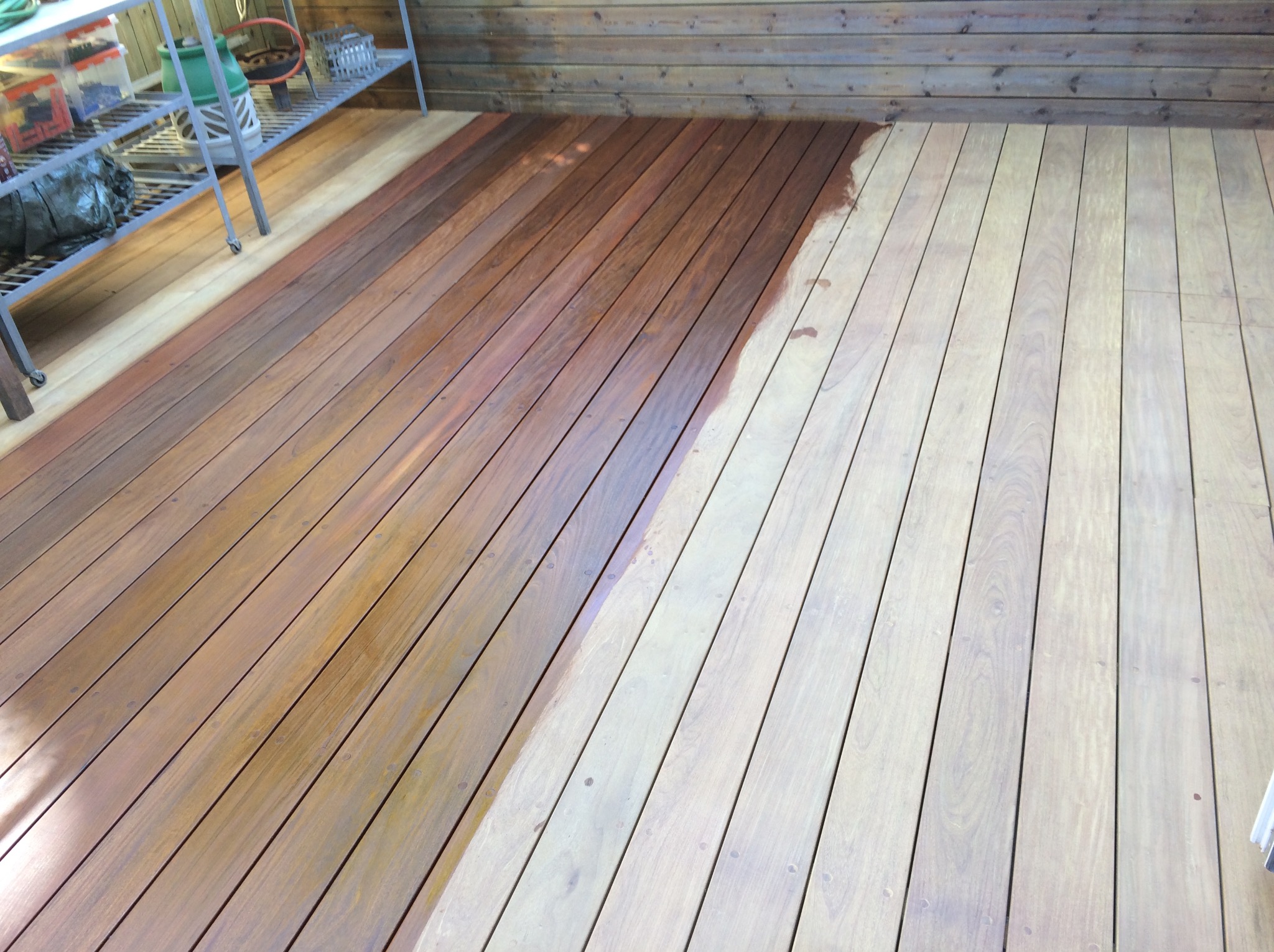 Drewniany taras, częściowo pokryty impregnatem w odcieniu brązu, widoczne różnice w kolorze drewna przed i po impregnacji, zarys stopy na świeżo nałożonej warstwie.