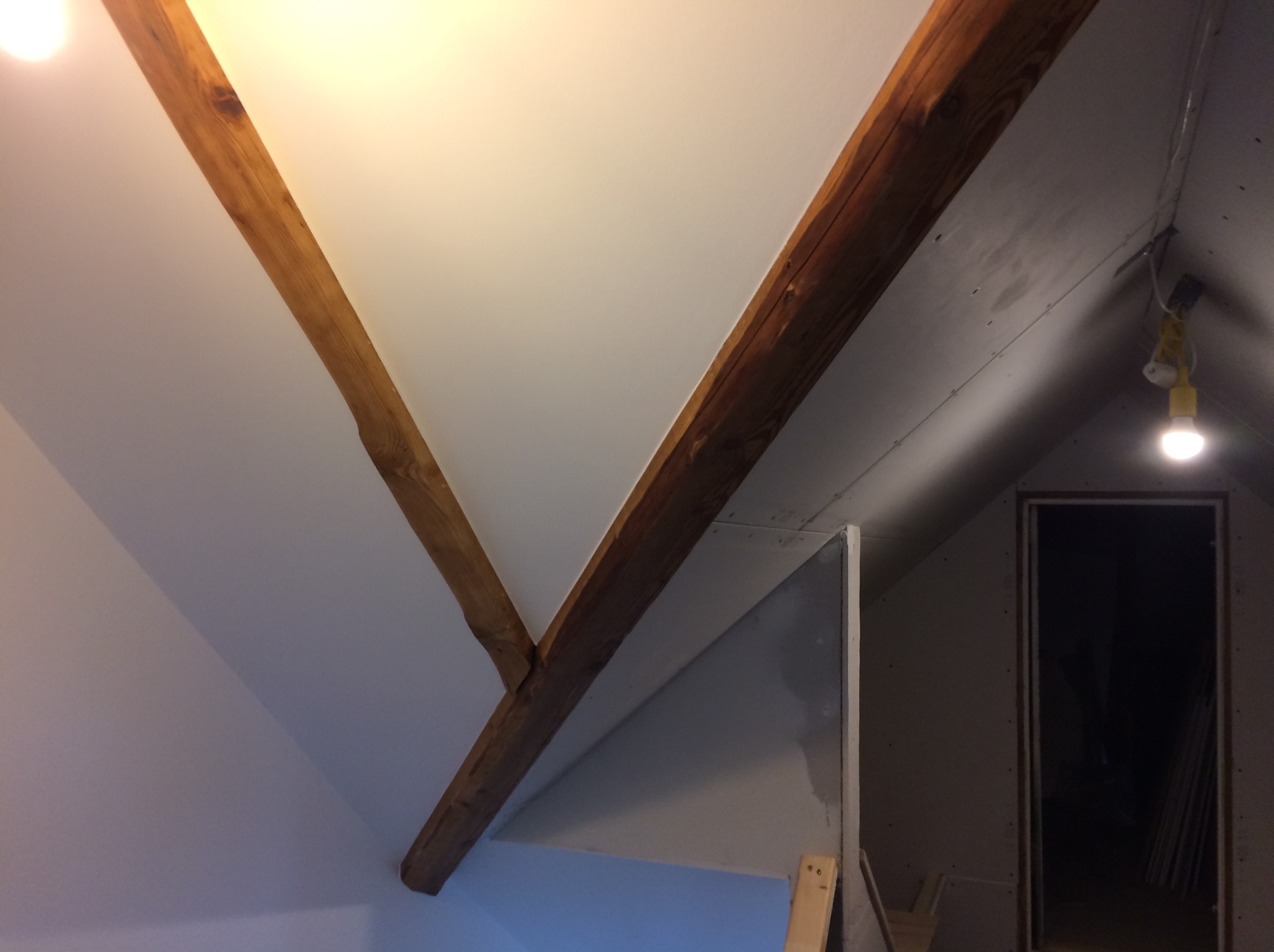 Poddasze w trakcie adaptacji: widoczne drewniane belki stropowe, fragment ściany z płyty karton gips i surowa ściana, wisząca lampa budowlana.