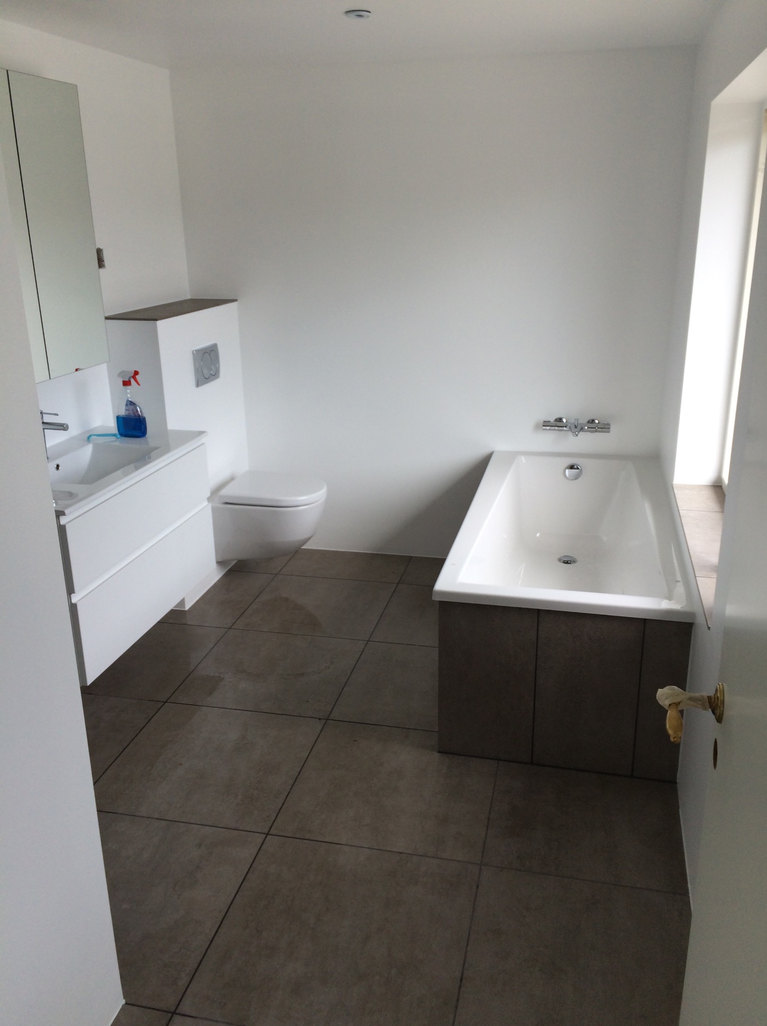 Nowoczesna łazienka z białymi ścianami, brązową podłogą z płytek, białą umywalką z szafką, toaletą podwieszaną i białą wanną z zabudową z ciemnego drewna.