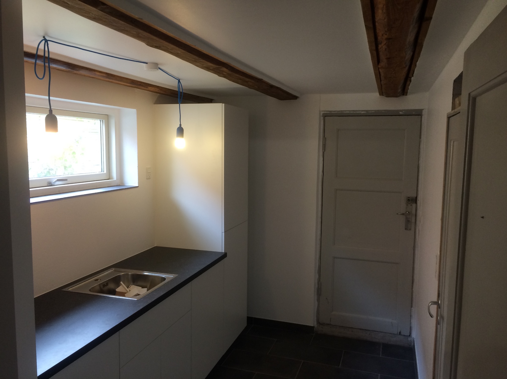 Wnętrze łazienki z widocznym oknem, umywalką w blacie, białymi szafkami, ciemną podłogą, belkami stropowymi i dwoma lampami z niebieskimi kablami.