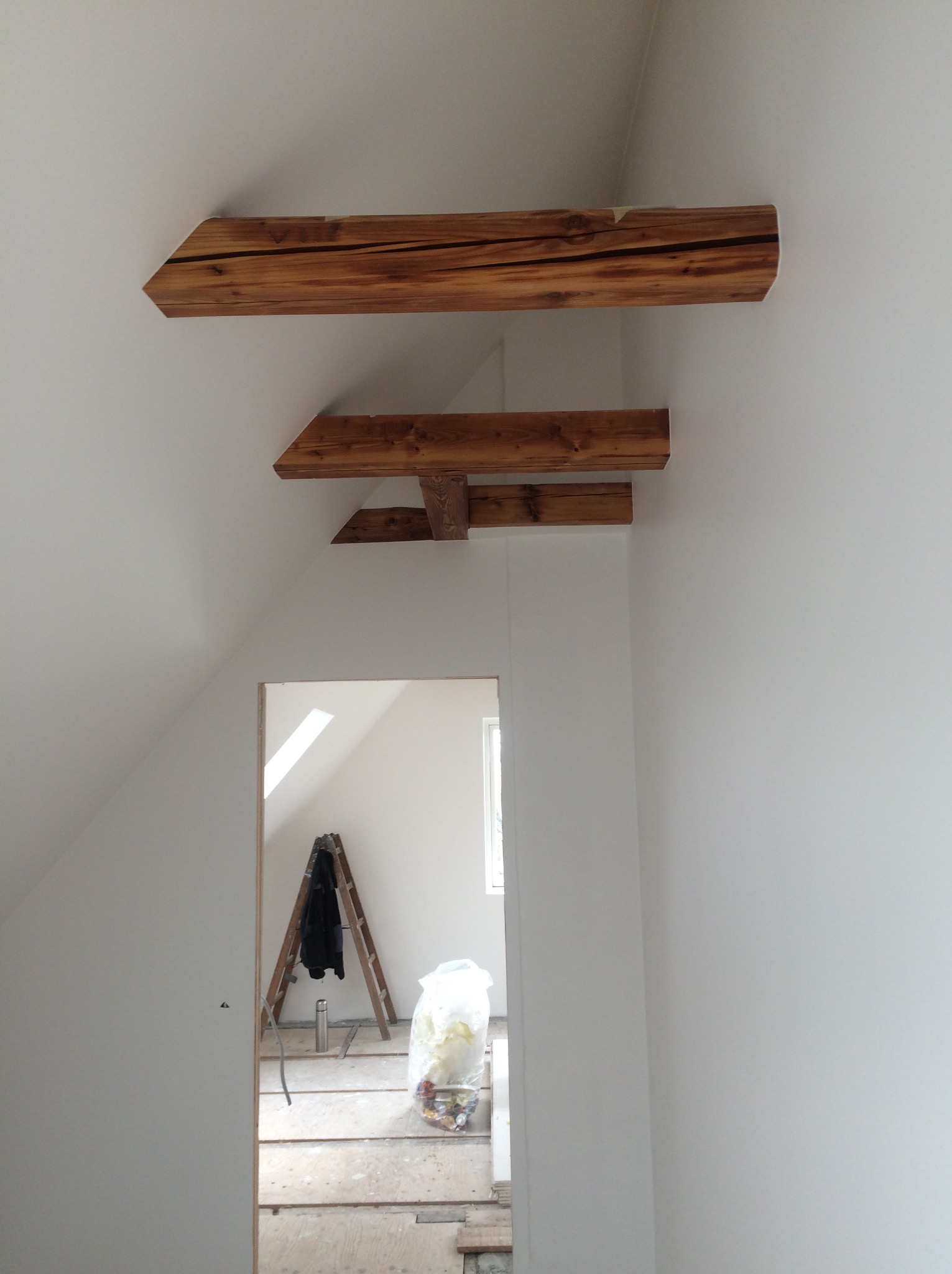 Poddasze w trakcie wykańczania: widoczne drewniane belki stropowe, białe ściany, drabina oparta o ścianę w pokoju obok oraz worki z odpadami budowlanymi.