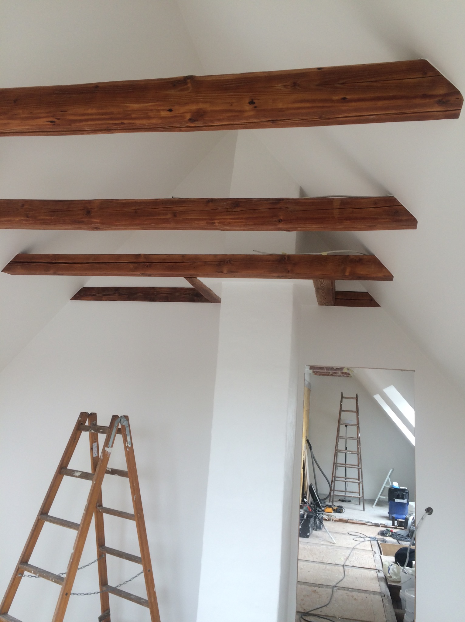 Poddasze po malowaniu na biało, widoczne drewniane belki stropowe, drabina malarska i sprzęt budowlany w sąsiednim pomieszczeniu