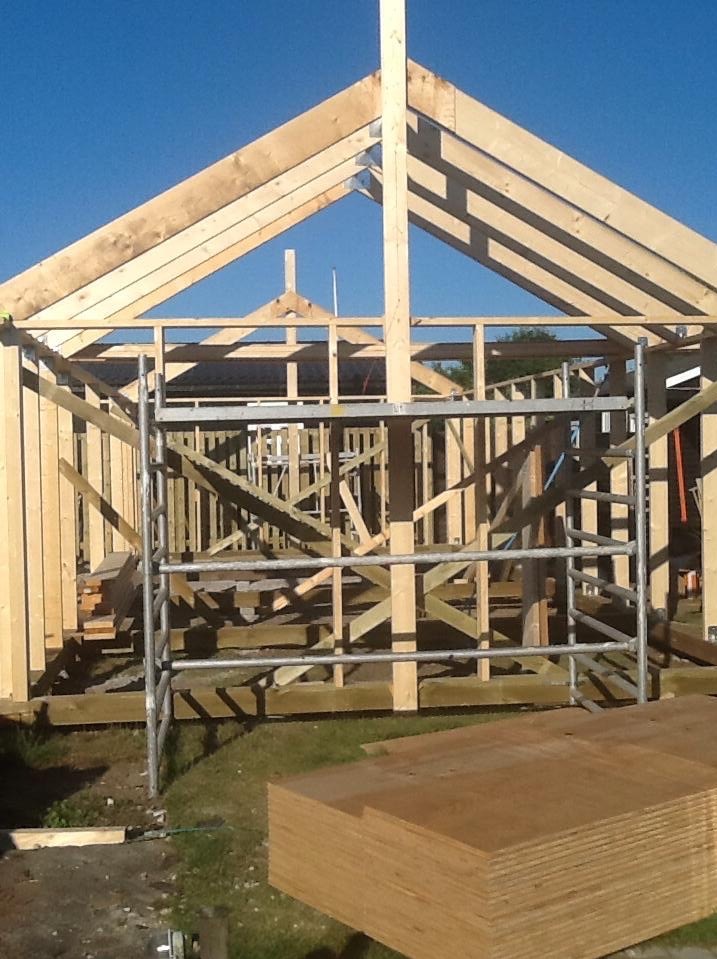 Konstrukcja drewniana szkieletu budynku w trakcie budowy, widoczne elementy więźby dachowej, ściany w surowym stanie, rusztowanie i stos płyt wiórowych na trawie.